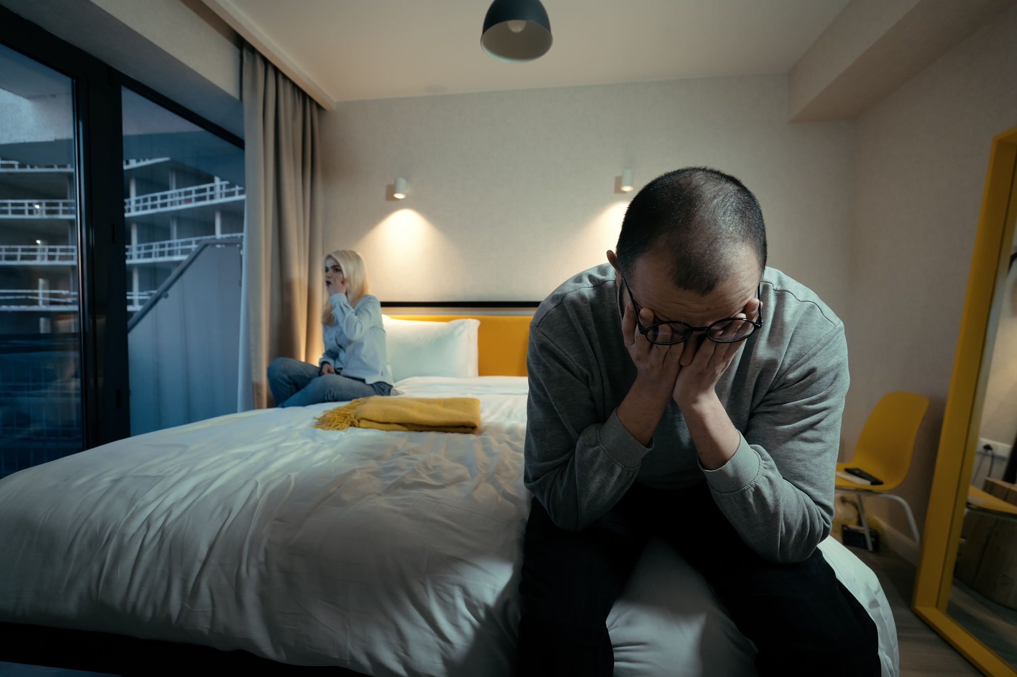 A man sits on the edge of a bed with his head in his hands, appearing distressed. A woman sits in the background on the bed, looking out the window. The room has modern décor, with large windows, yellow accents, and a neatly made bed.