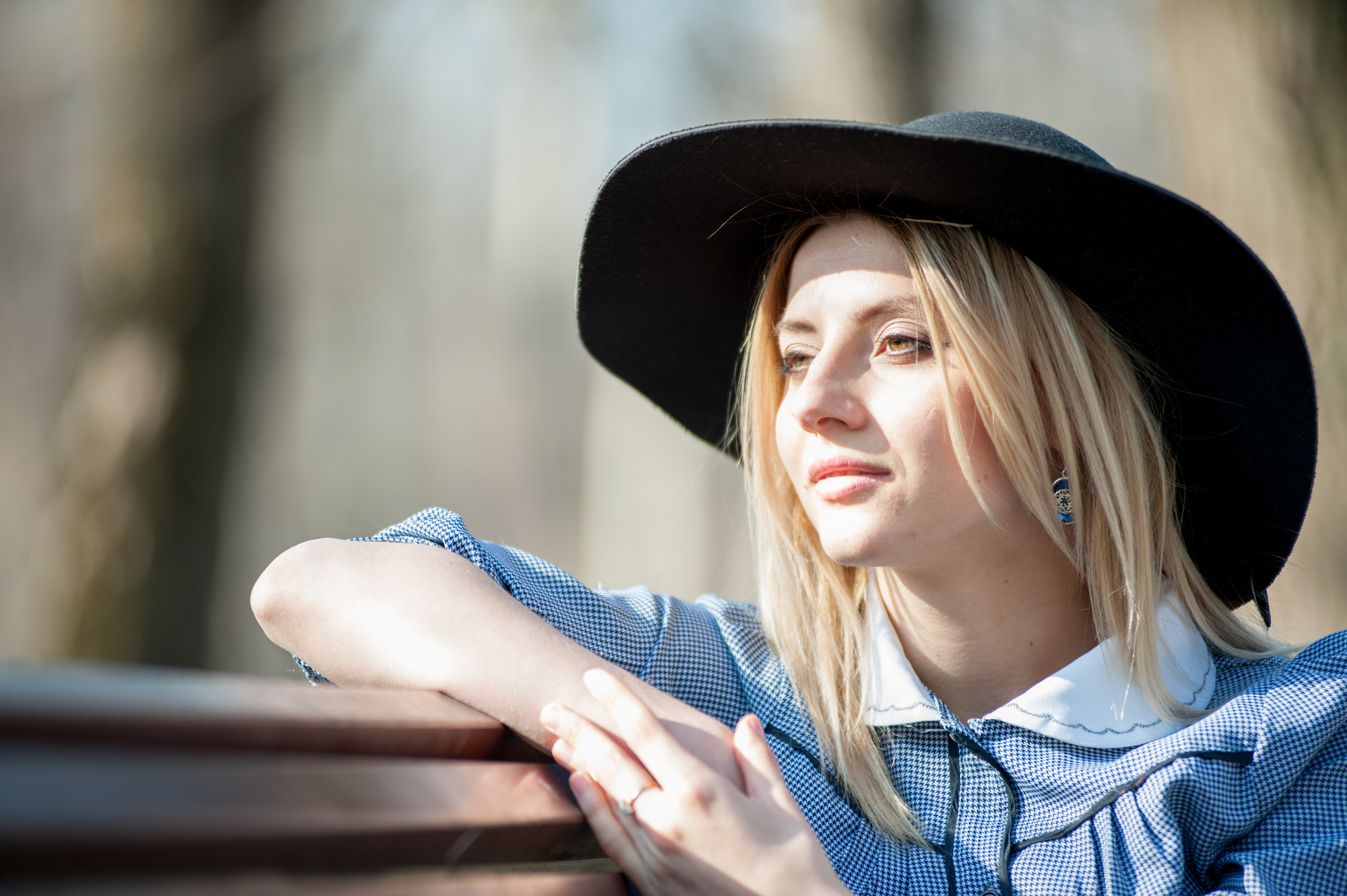 A woman wearing a large, black sunhat and a blue checkered dress with a white collar leans on the back of a bench. She looks forward thoughtfully, with sunlight illuminating her face against a blurred, natural background.