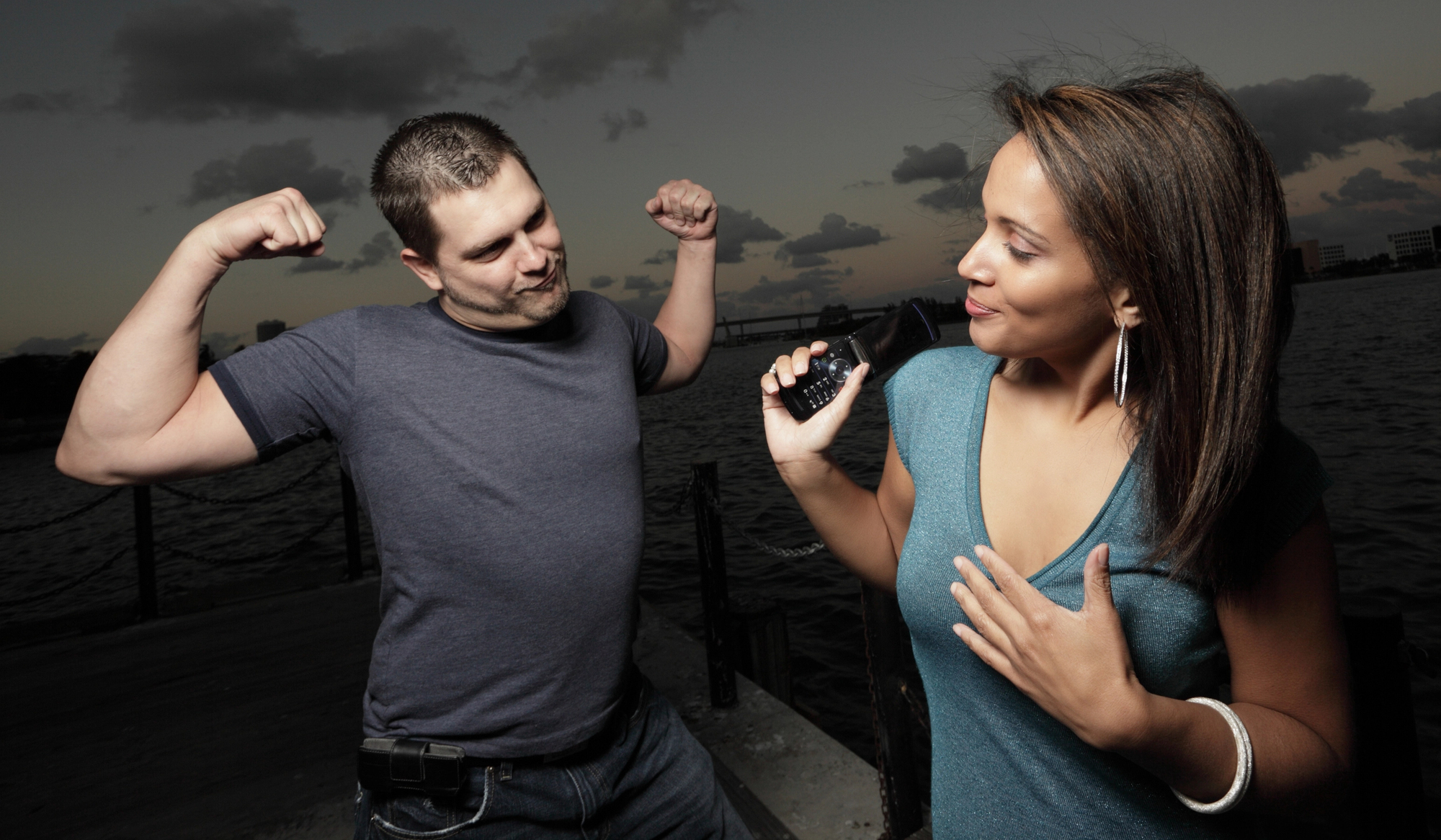 A man in a gray t-shirt flexes his biceps enthusiastically while a smiling woman in a teal top holds a microphone towards him. They are outdoors near water with a cloudy sky in the background.