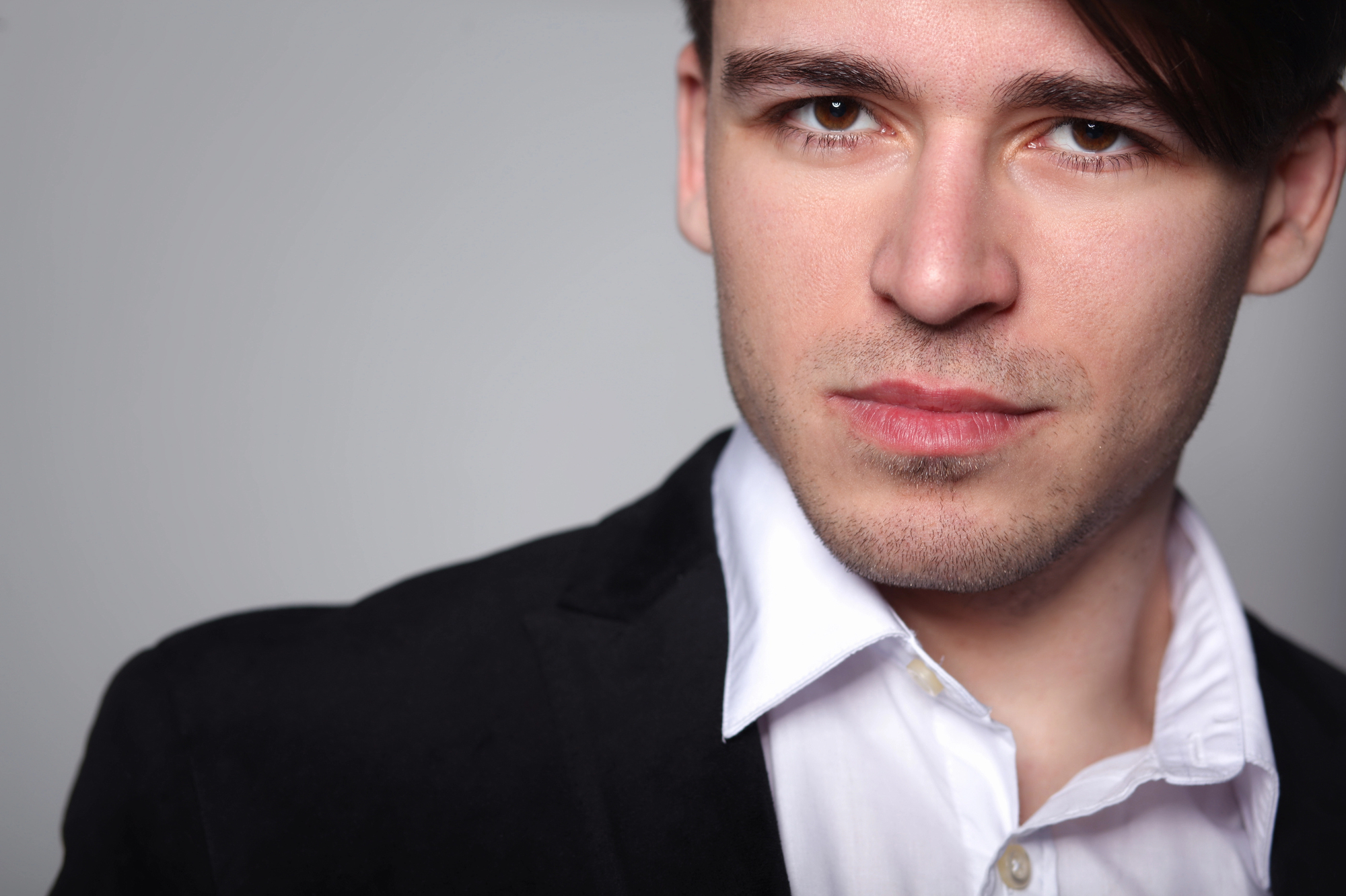 A young man with light skin and short dark hair gazes at the camera with a slight smile. He is wearing a white dress shirt under a black suit jacket. The background is plain and gray.