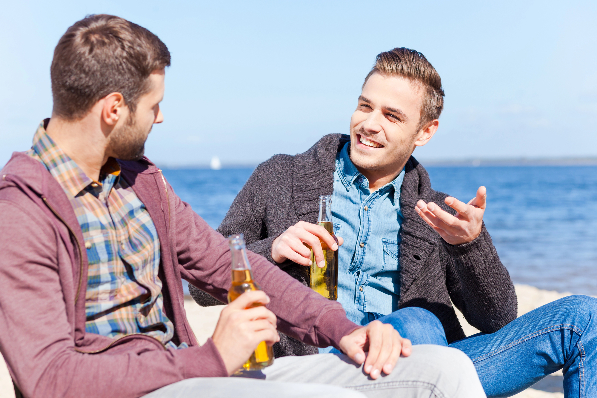 Two men sitting on a sandy beach, each holding a beer bottle. They are engaged in a lively conversation, with the man on the right gesturing animatedly. Both are casually dressed in layered clothing. The sea is visible in the background under a clear blue sky.