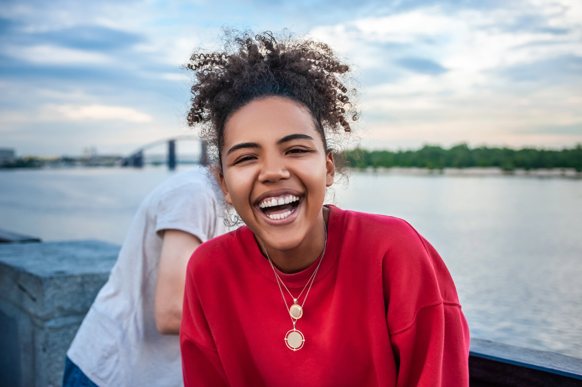 A young woman with curly hair, wearing a red sweatshirt and layered necklaces, smiles brightly at the camera. She stands outdoors near water with a bridge and greenery in the background. A part of another person is visible behind her.