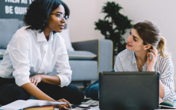 Two women sit on the floor in front of an open laptop, engaged in discussion. One has braided hair and wears glasses and a white shirt; the other has light hair tied back and wears a striped shirt. An open book lies in front of them, with a gray couch in the background.