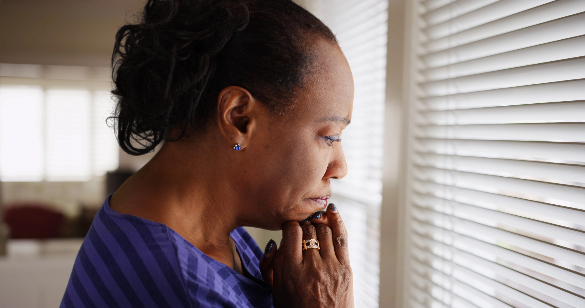 A woman with a thoughtful expression gazes out of a window, partially covered by venetian blinds. She has her hands near her chin and is wearing a blue striped shirt and earrings. The room is softly lit with natural light filtering through the blinds.