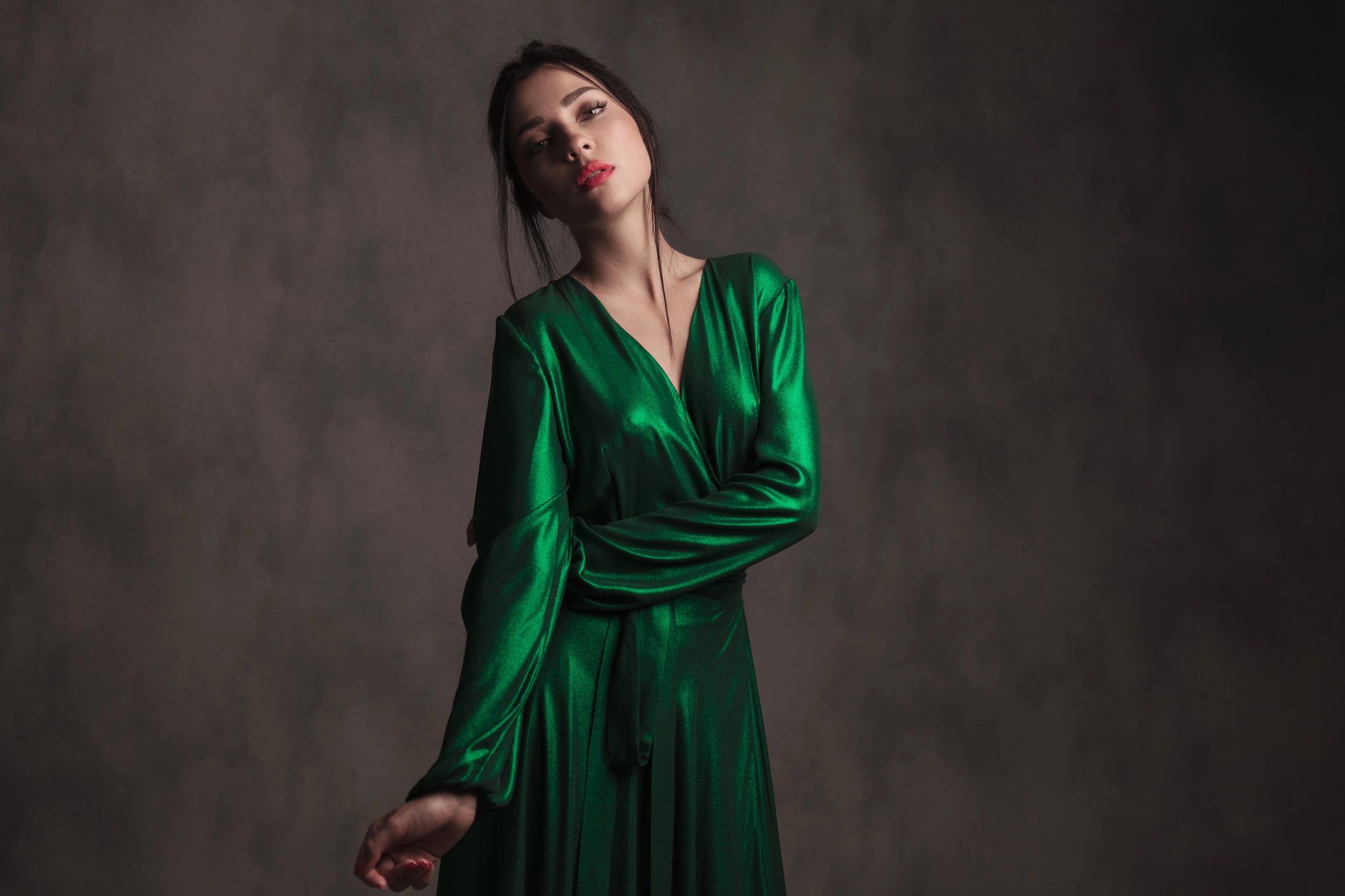 A person stands in front of a dark, textured background, wearing a shiny green dress. The lighting creates shadows, giving a dramatic effect. The person’s head is slightly tilted, and their arms are crossed, adding a sense of elegance to the pose.