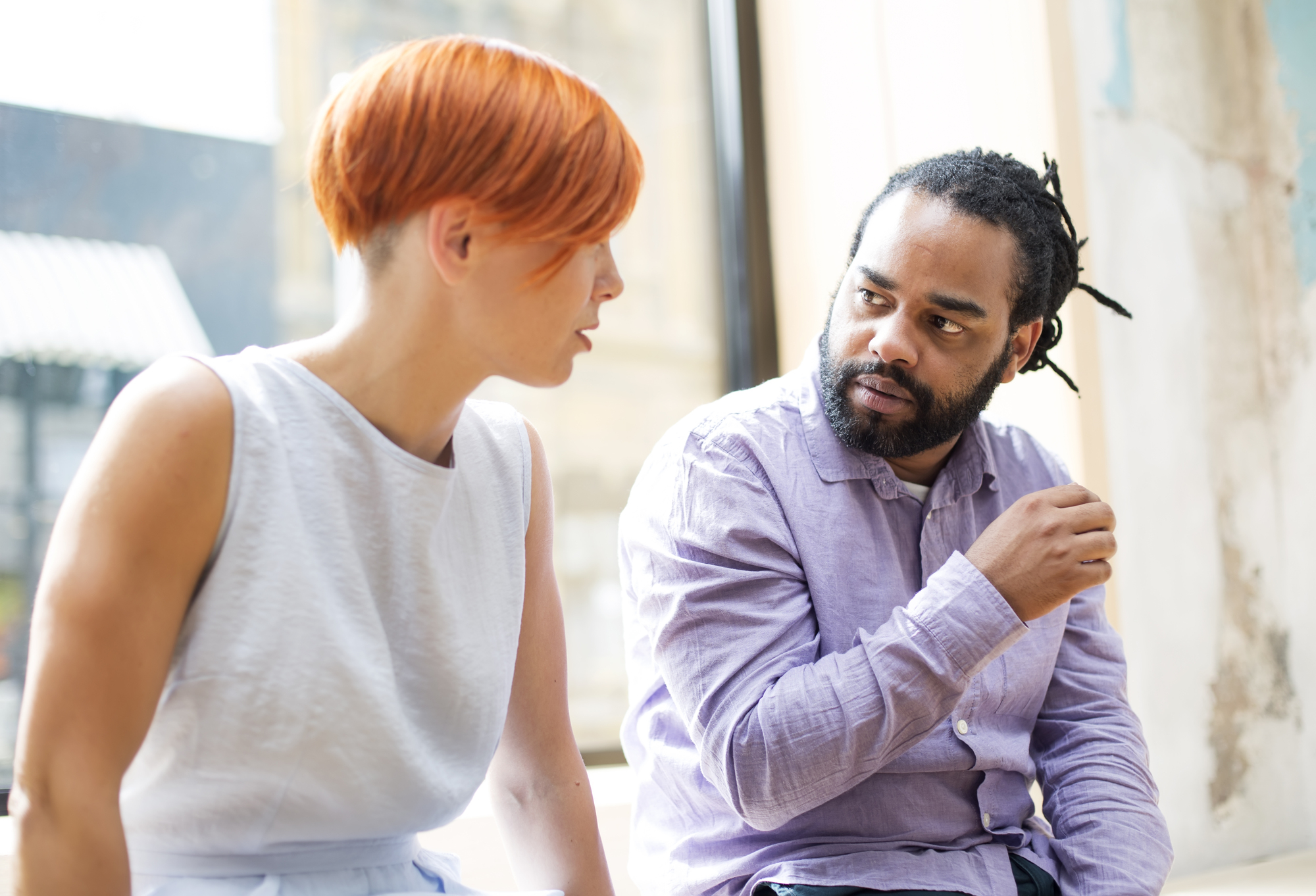 A woman with short red hair is engaged in a serious conversation with a bearded man with dreadlocks who is gesturing with his hand. They are indoors, with a large window in the background providing natural light. Both are focused on each other.