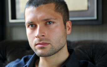 A person with short brown hair and a trimmed beard gazes pensively into the distance while seated indoors. They are wearing a dark-colored shirt. The background is blurred, focusing attention on the contemplative expression on their face.