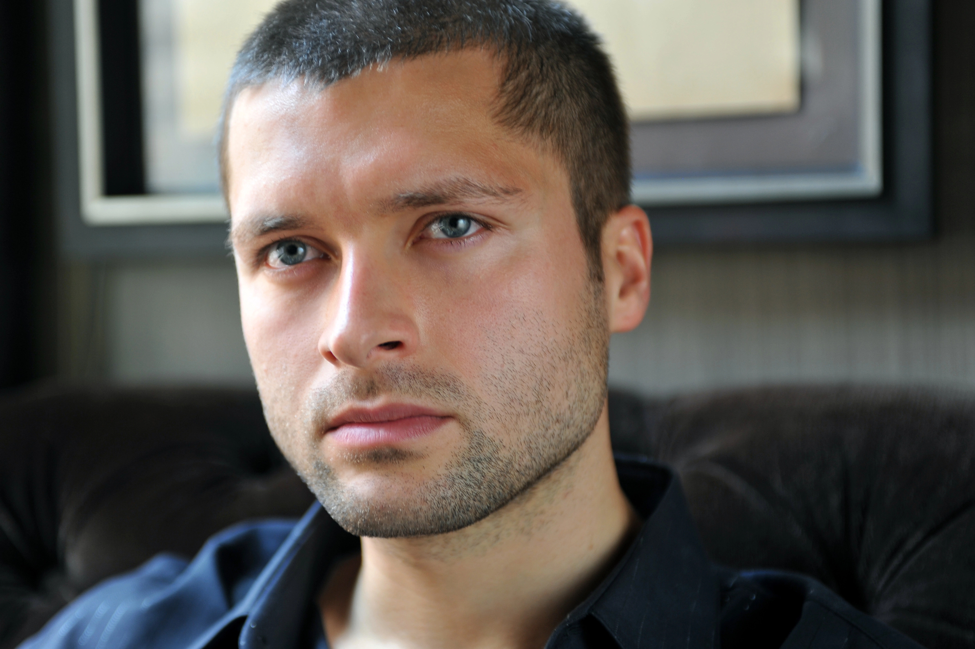 A person with short brown hair and a trimmed beard gazes pensively into the distance while seated indoors. They are wearing a dark-colored shirt. The background is blurred, focusing attention on the contemplative expression on their face.