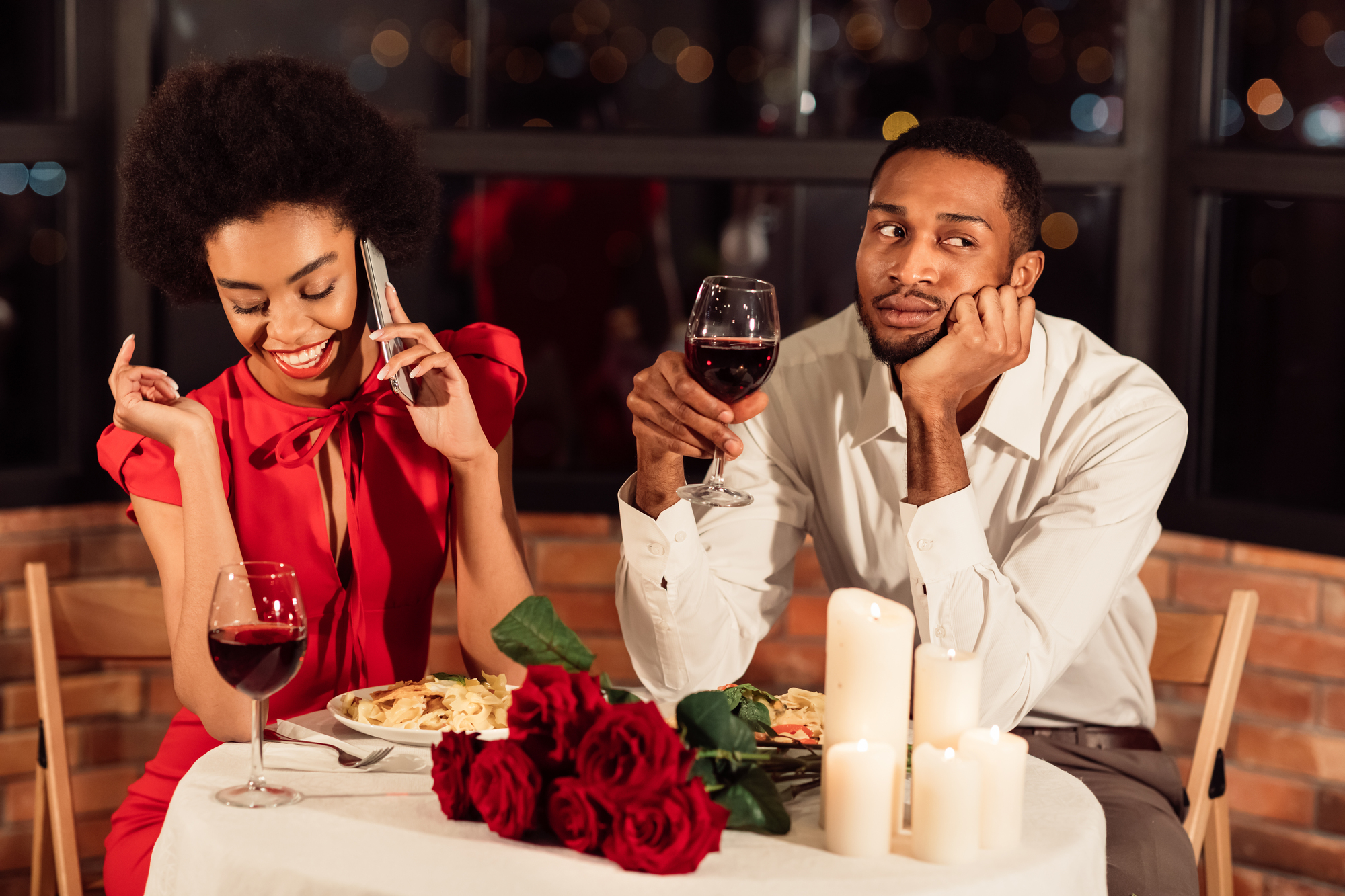 A woman in a red dress smiles while talking on her cellphone during a candlelit dinner. A man in a white shirt sits beside her, looking bored and holding a glass of red wine. A bouquet of red roses and more wine are on the table, set against a city night backdrop.