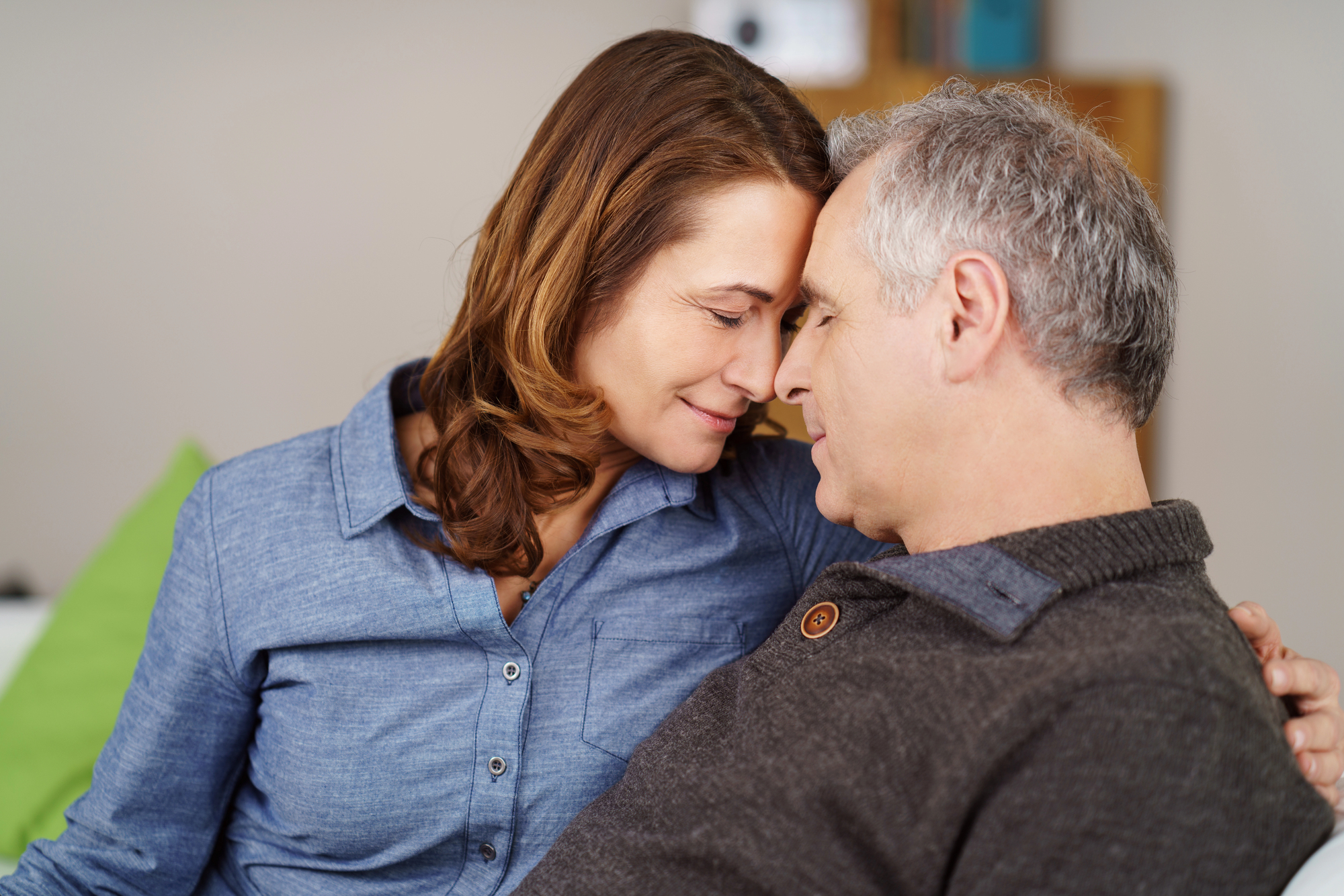 A woman and a man are sitting closely together, smiling with their foreheads touching. The woman has brown hair and is wearing a blue shirt, while the man has gray hair and is wearing a black sweater. They both appear to be happy and content.