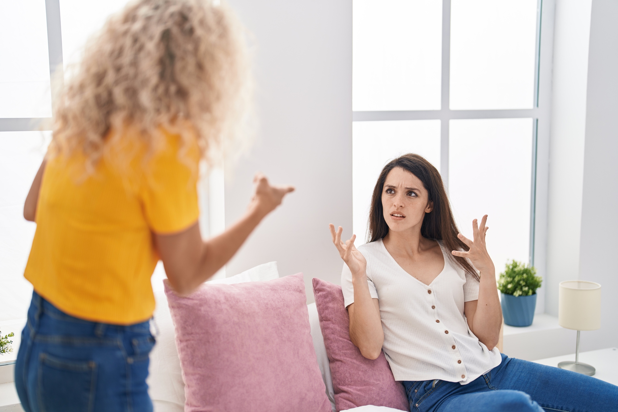 Two women are in a brightly lit room. One, with curly blonde hair and wearing a yellow shirt, is standing and pointing, appearing angry. The other, with straight dark hair and wearing a white shirt, is sitting on a bed looking confused and defensive.