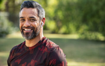 A smiling man with salt-and-pepper hair and a short beard is standing outdoors. He is wearing a red and black camouflage shirt and has white wireless earphones in his ears. The background is blurred, showing trees and greenery.