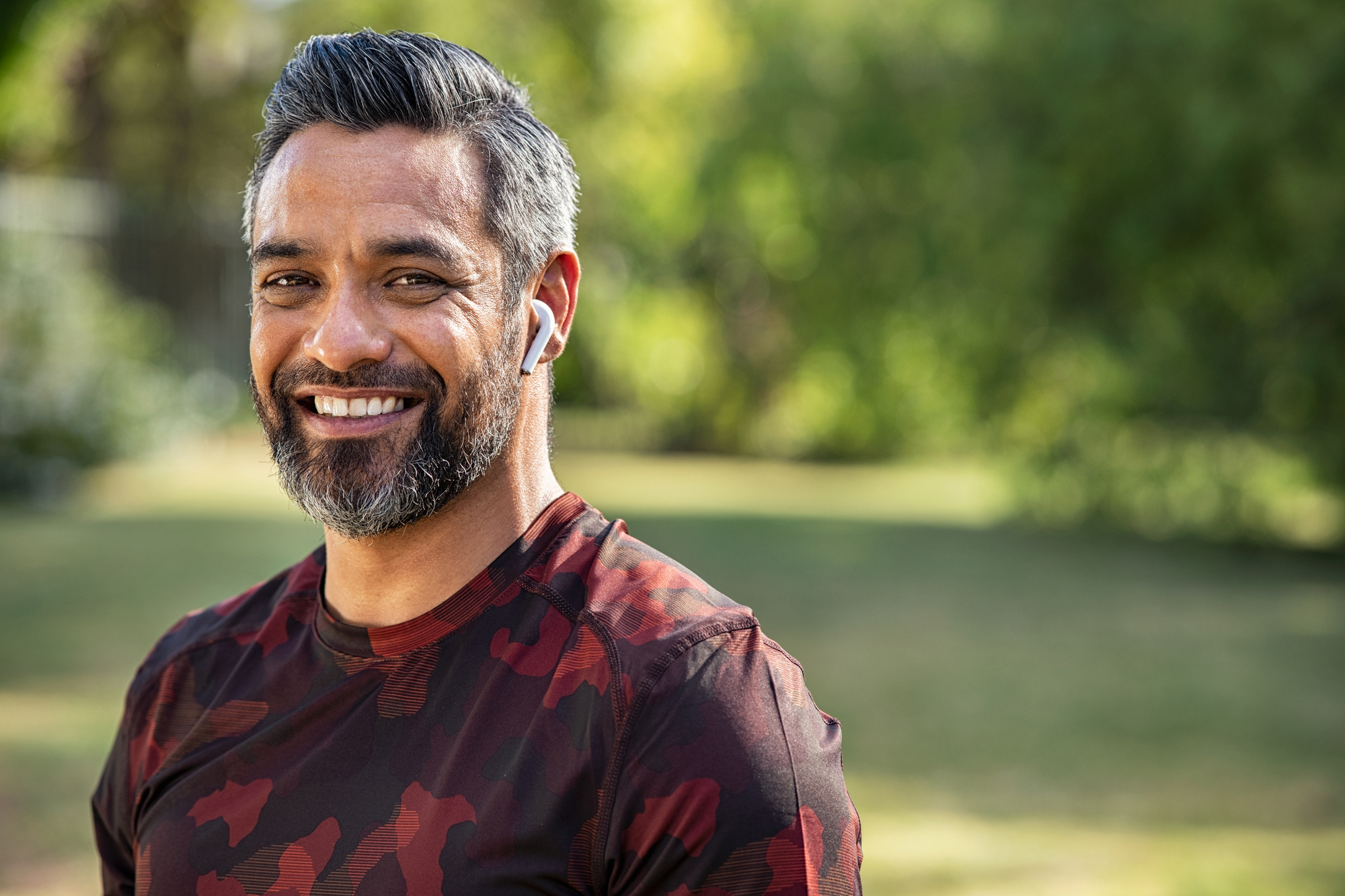 A smiling man with salt-and-pepper hair and a short beard is standing outdoors. He is wearing a red and black camouflage shirt and has white wireless earphones in his ears. The background is blurred, showing trees and greenery.