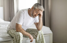 A middle-aged man with gray hair, wearing a white t-shirt and plaid pajama pants, sits on the edge of a bed with his head resting in one hand, appearing troubled or stressed. The room is softly lit with neutral-colored curtains in the background.