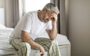 A middle-aged man with gray hair, wearing a white t-shirt and plaid pajama pants, sits on the edge of a bed with his head resting in one hand, appearing troubled or stressed. The room is softly lit with neutral-colored curtains in the background.