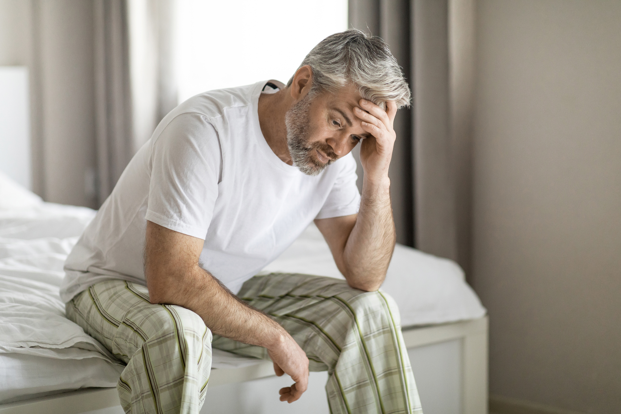 A middle-aged man with gray hair, wearing a white t-shirt and plaid pajama pants, sits on the edge of a bed with his head resting in one hand, appearing troubled or stressed. The room is softly lit with neutral-colored curtains in the background.