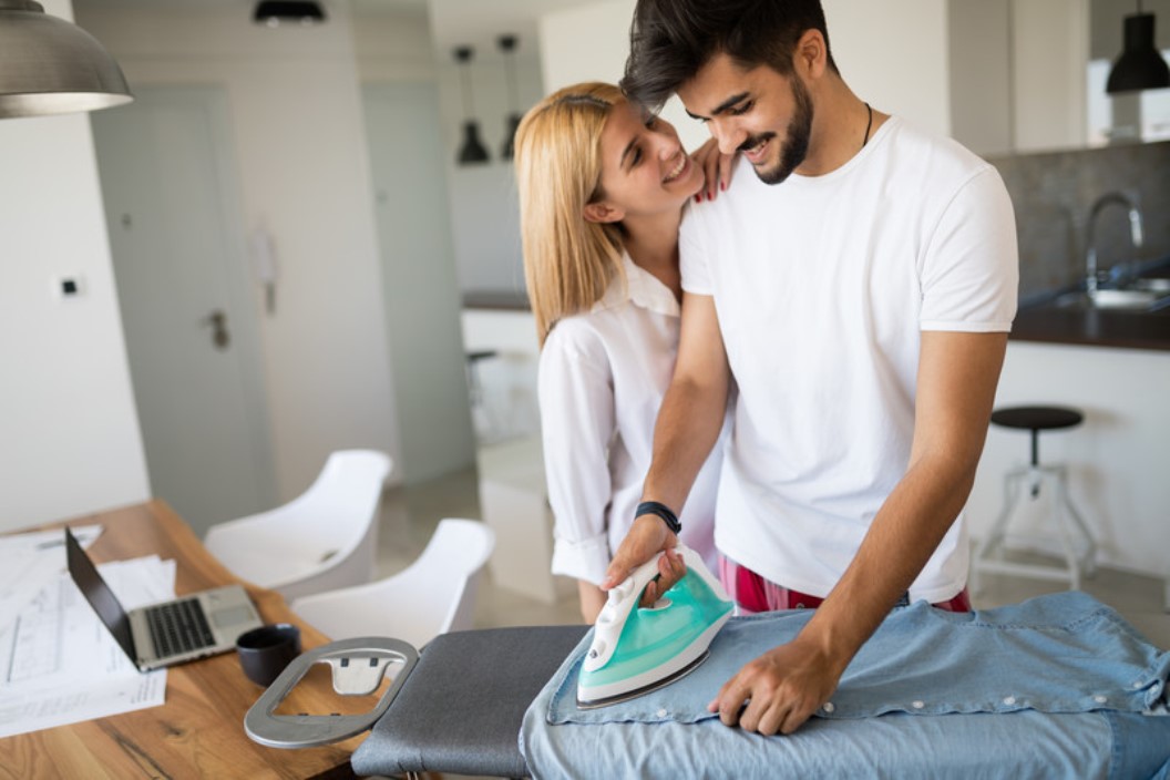 A smiling man is ironing a pair of jeans on an ironing board in a modern kitchen while a woman, also smiling, stands next to him with her arm around his shoulder. A laptop, coffee cup, and papers are on the table in the background.