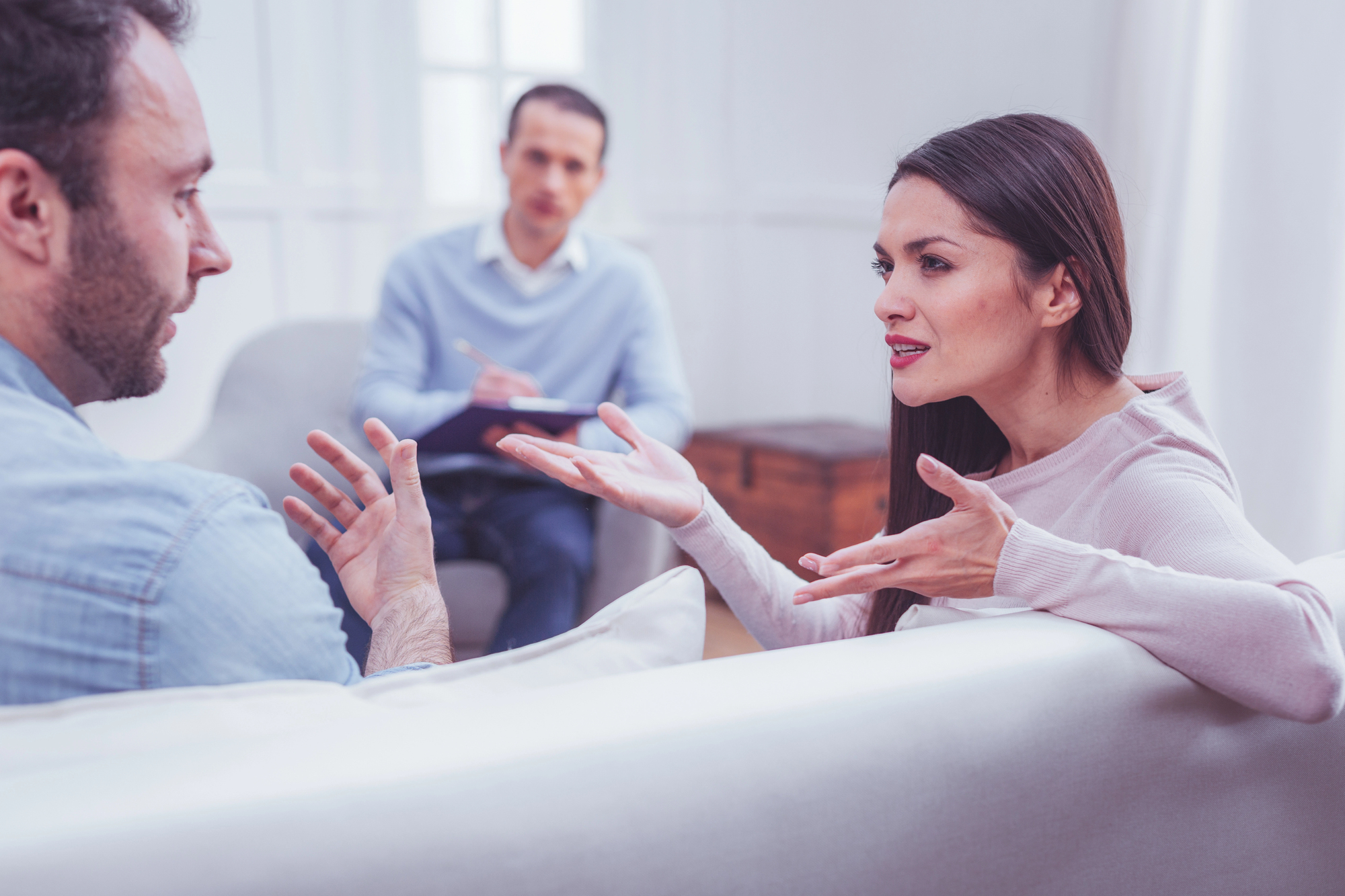 A couple sits on a couch facing each other, engaged in a heated discussion, gesturing with their hands. A therapist, holding a clipboard and pen, sits in a chair in the background, listening attentively. The scene appears to be a counseling session.