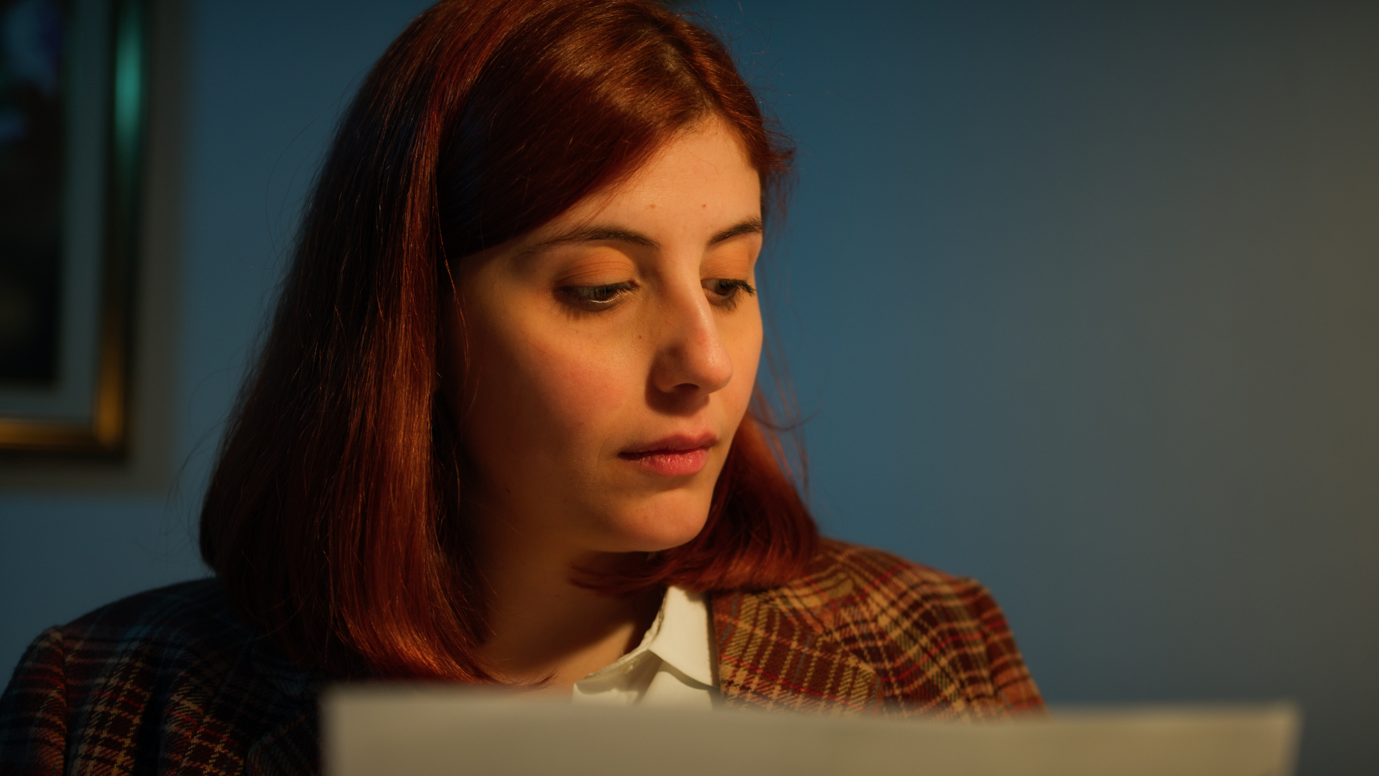 A person with shoulder-length reddish hair is looking slightly downward, with a neutral expression, illuminated by warm lighting. They are wearing a checkered blazer over a collared shirt and appear to be reading or examining something out of frame.