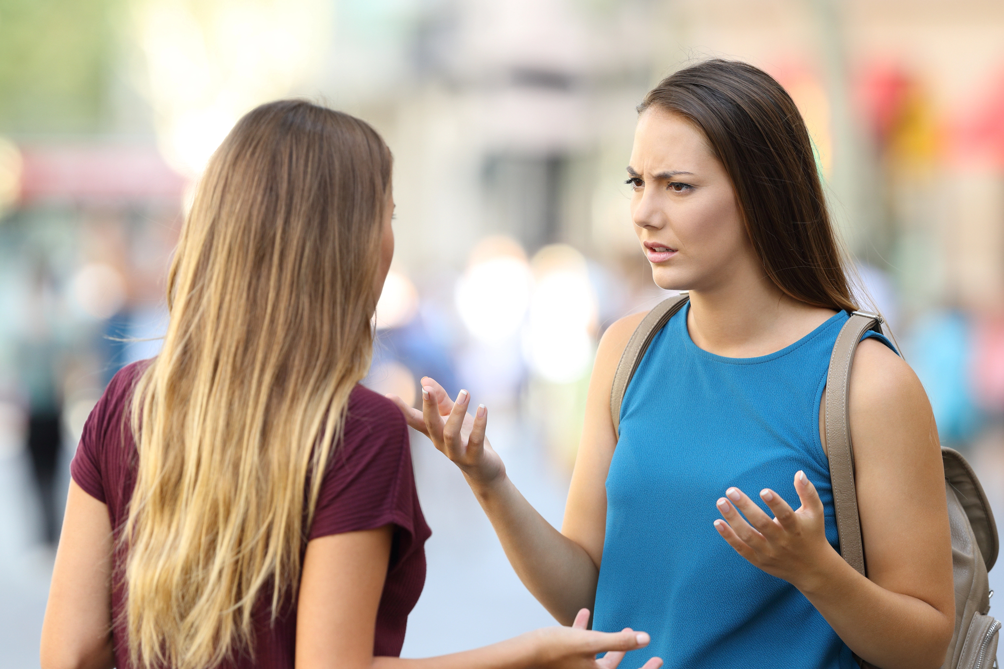 Two women are having a serious conversation outdoors. One woman with her back to the camera has long blonde hair and is wearing a maroon shirt. The other woman with long brown hair and wearing a blue tank top has a concerned expression and gesturing with her hands.