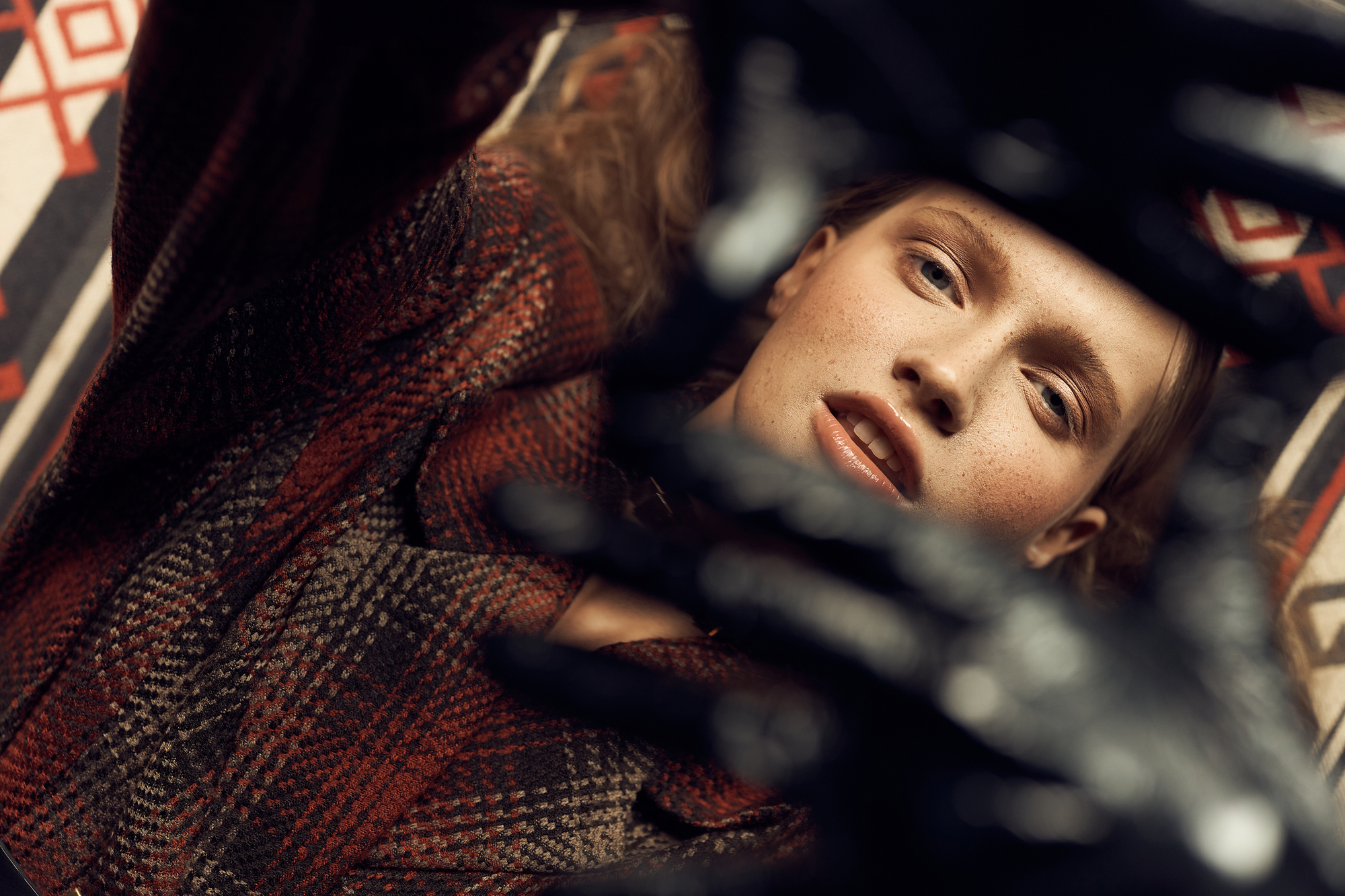 A person with light skin and freckles, wearing a red and black checkered coat, lies on a patterned fabric while looking up into the camera. Part of the image is obscured by a black object in the foreground.