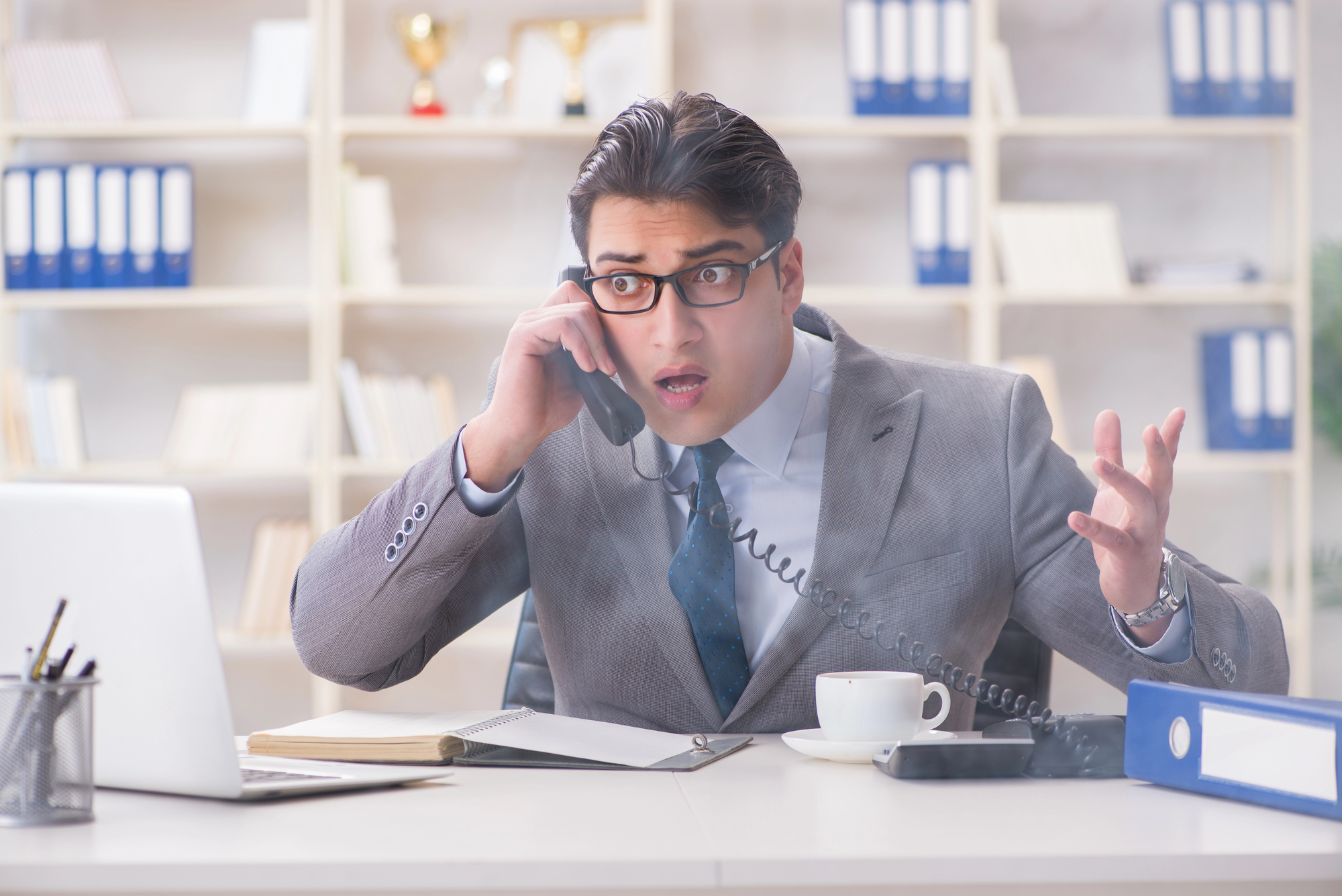 A man wearing glasses and a suit is sitting at a desk in an office, holding a phone to his ear with an expression of surprise or frustration. There are bookshelves with binders and trophies in the background, and a laptop, notebook, and coffee cup on the desk.