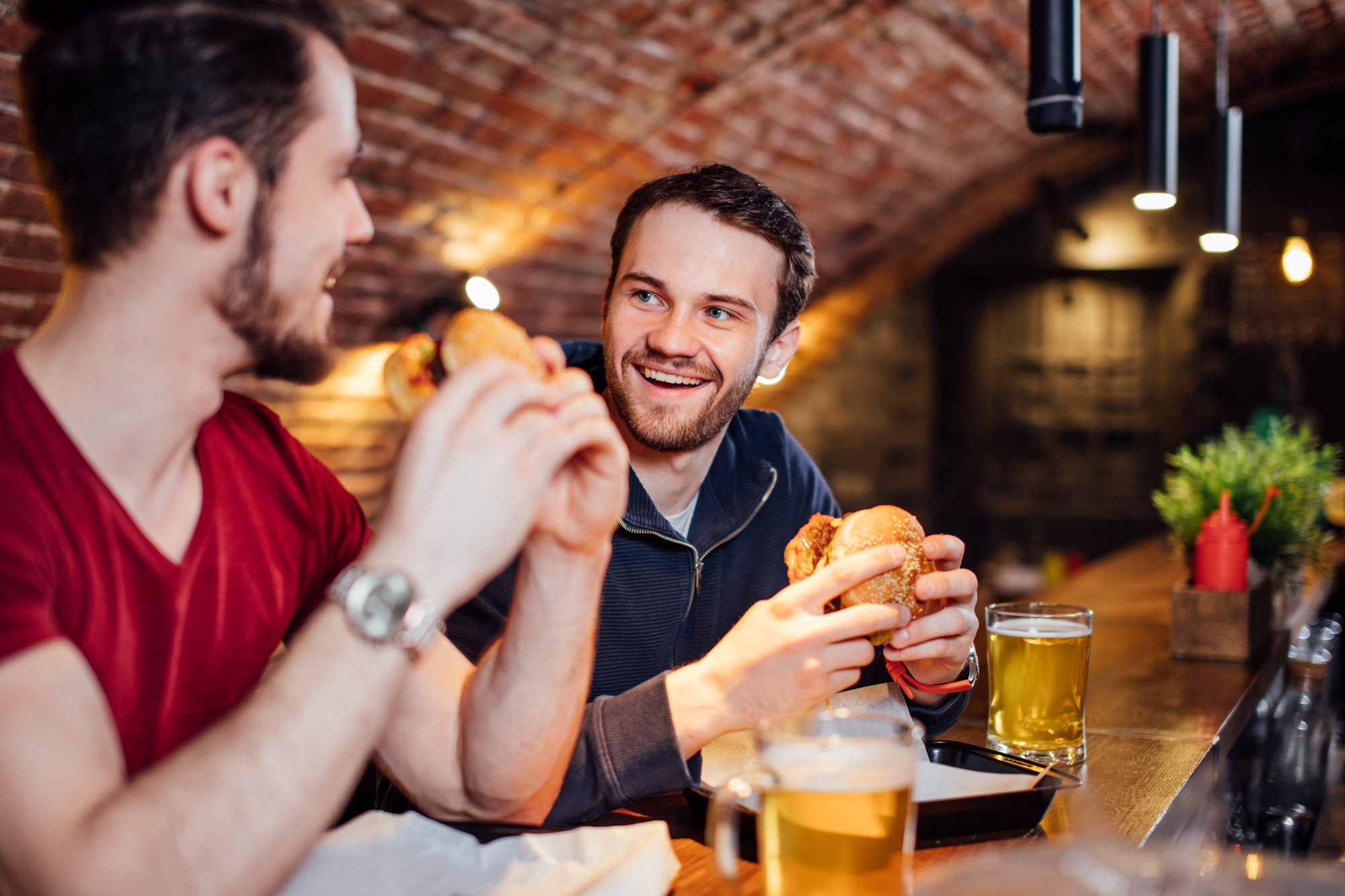 Two friends sit at a wooden bar in a cozy, dimly-lit pub with brick arch ceilings. They are smiling and laughing while holding burgers. A glass of beer is on the counter in front of each of them. Decorative plants and condiments are also on the countertop.