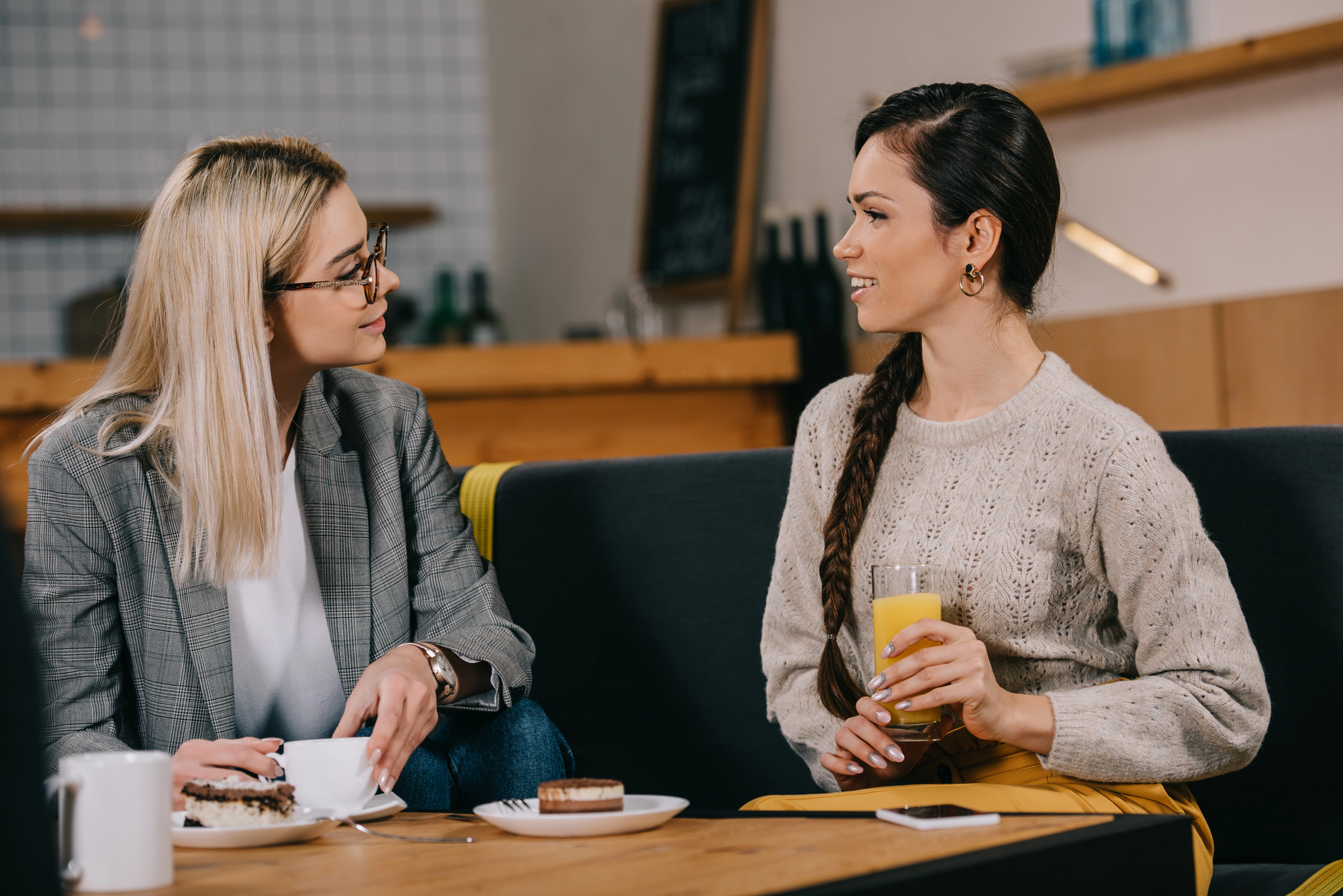 Two women sitting at a cafe table, engaged in conversation. One woman, wearing glasses and a grey blazer, holds a cup of coffee and a pastry. The other woman, in a beige sweater with a long braid, holds a glass of juice. They appear to be enjoying a casual chat.