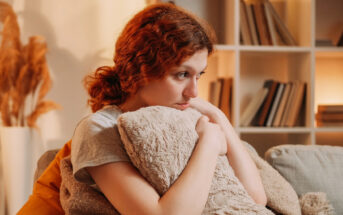 A young woman with curly red hair sits on a couch, hugging a fluffy pillow and looking pensive. She is surrounded by a cozy living room with bookshelves and warm lighting in the background, creating a comfortable and contemplative atmosphere.