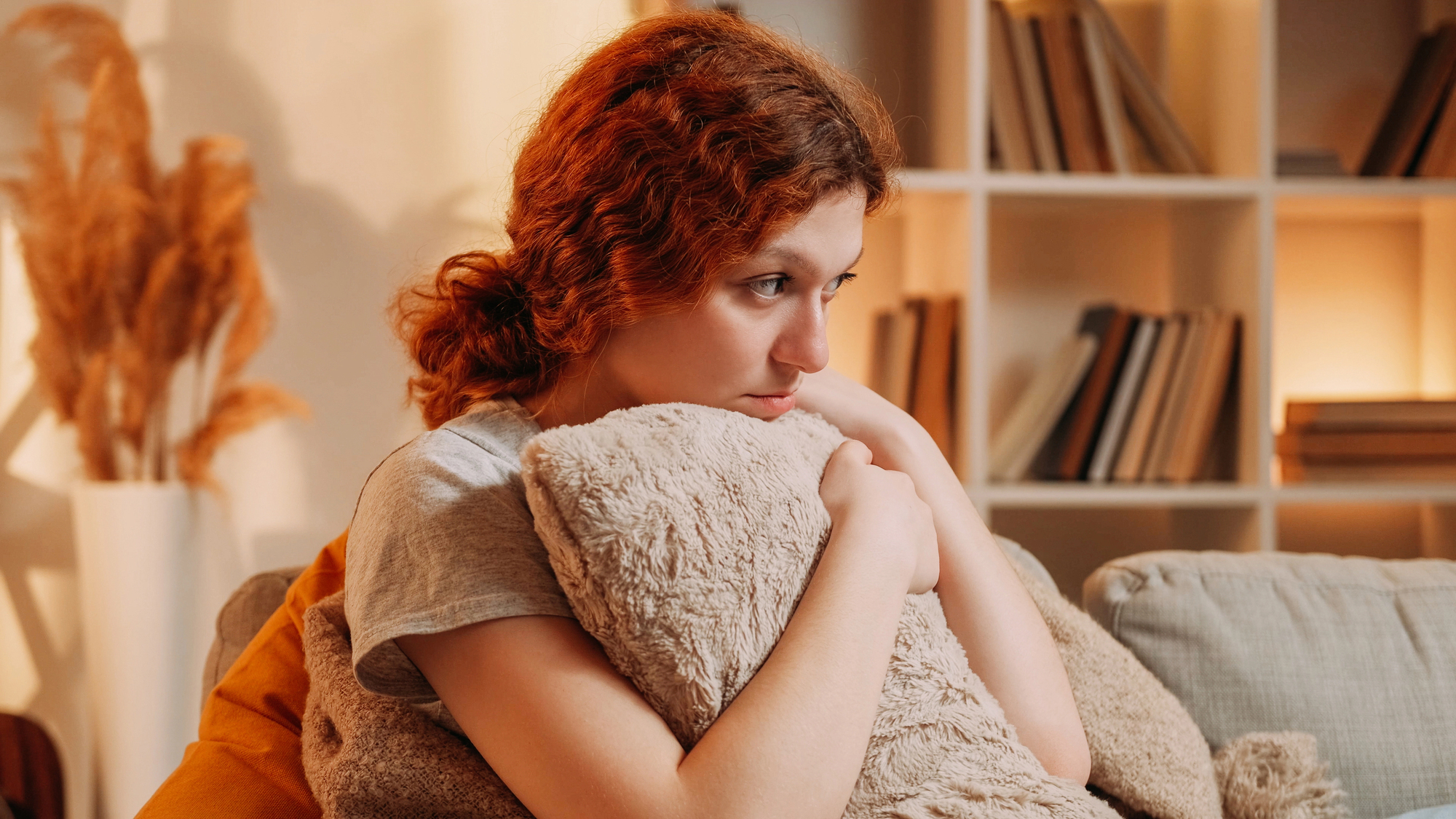 A young woman with curly red hair sits on a couch, hugging a fluffy pillow and looking pensive. She is surrounded by a cozy living room with bookshelves and warm lighting in the background, creating a comfortable and contemplative atmosphere.