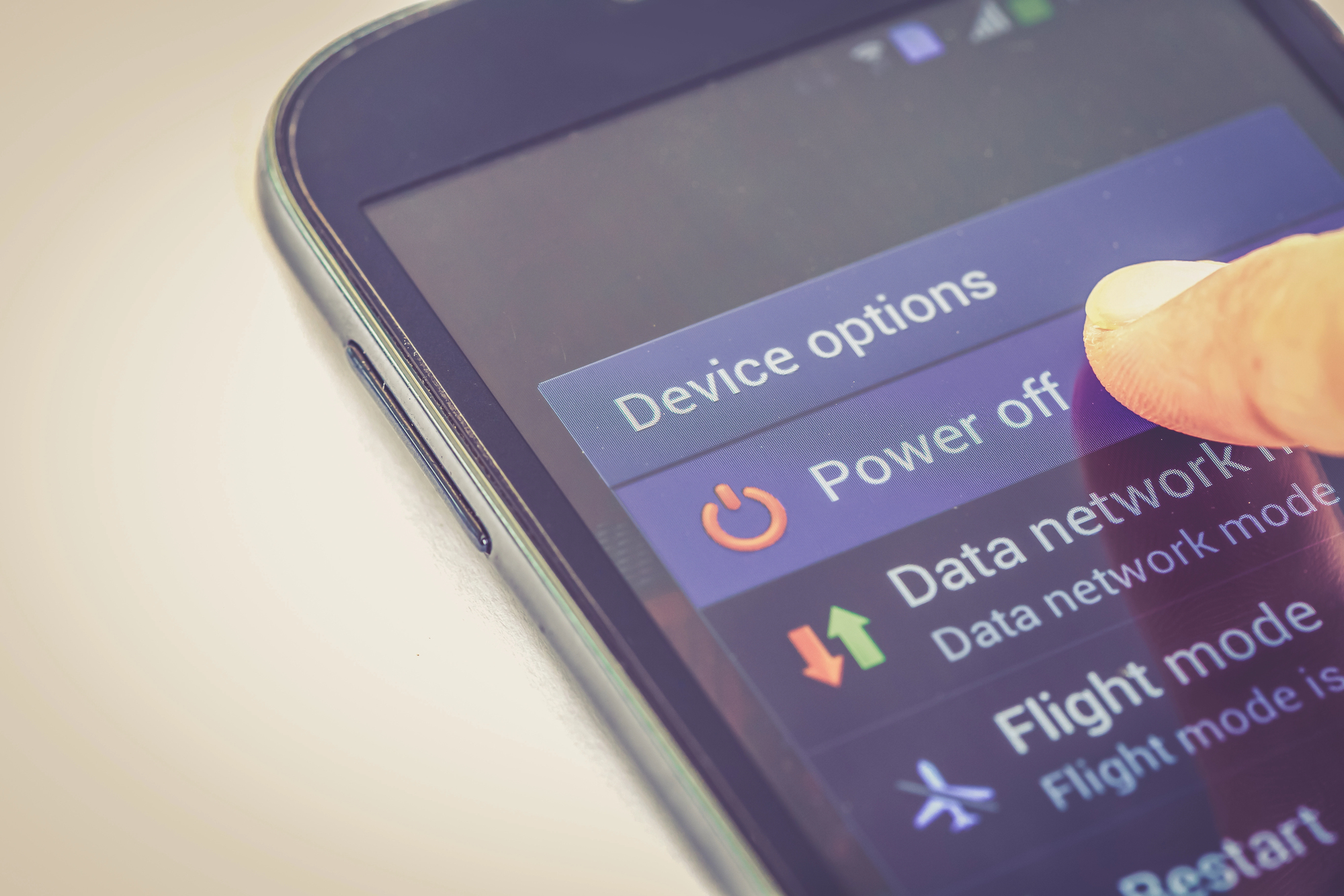 Close-up of a smartphone screen showing the "Device options" menu. A person's finger is about to select the "Power off" option. Other options like "Data network mode," "Flight mode," and "Restart" are also visible on the screen.