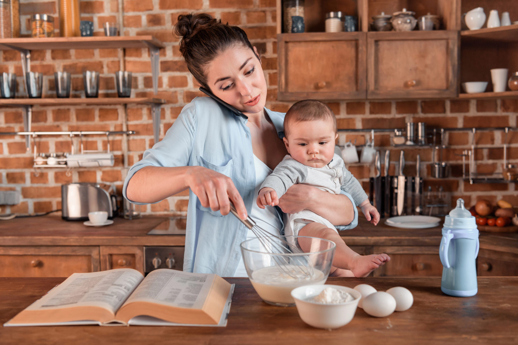 A woman multitasks in a kitchen by holding a baby in one arm while whisking ingredients in a bowl with the other hand. A cookbook is open on the counter next to eggs, flour, and a baby bottle. The background features wooden shelves, dishes, and kitchen utensils.