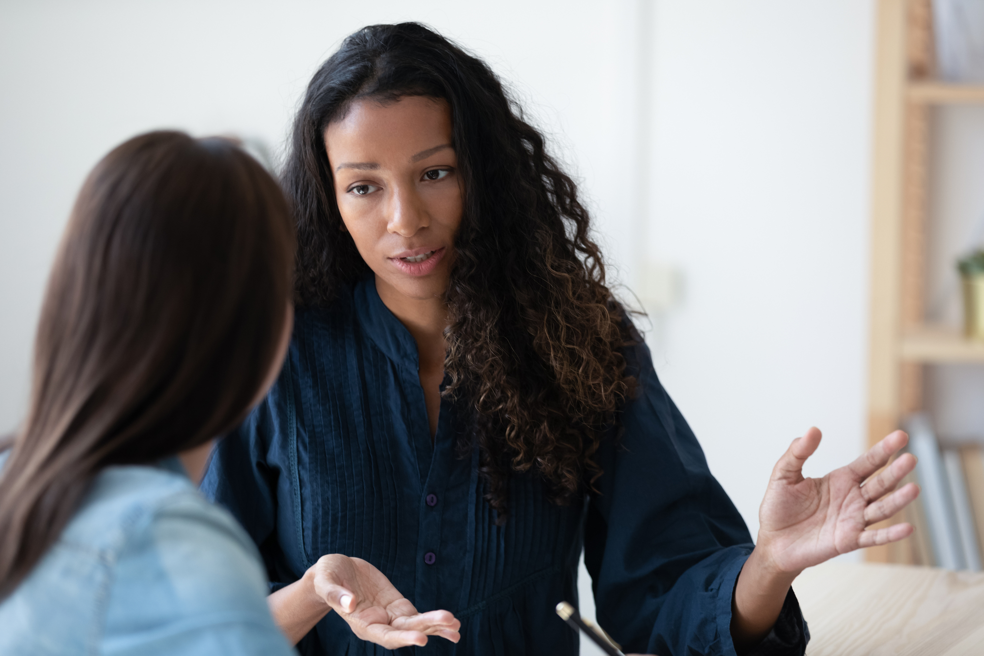 A woman with long curly hair and wearing a dark blue shirt gestures with her hands while speaking to another woman with straight hair, who is blurred in the foreground. They appear to be having a serious conversation in a bright, indoor setting.