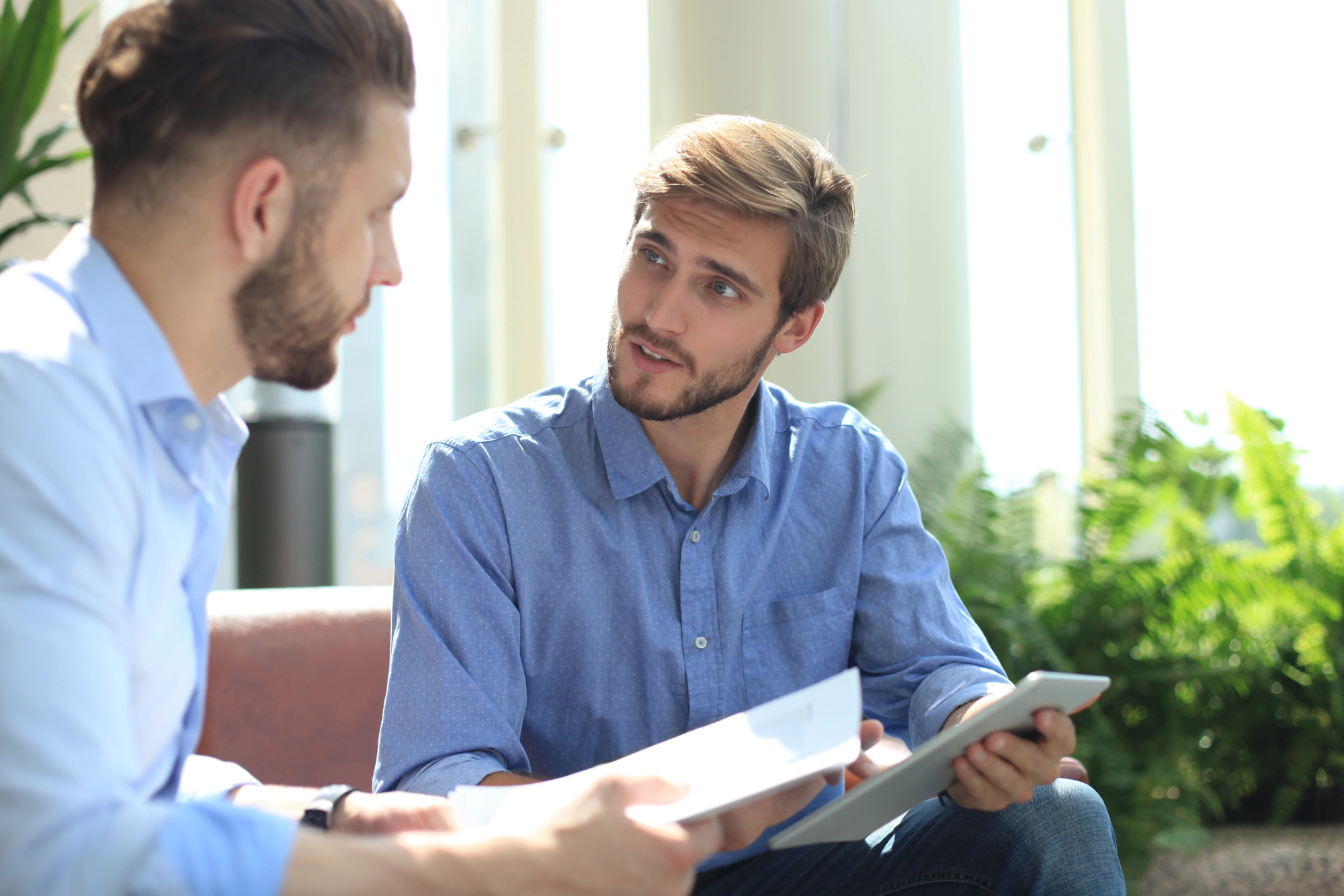 Two men are sitting and talking in a bright room with large windows. One man on the right holds a tablet and papers while looking at the man on the left, who gestures with his hand. Both wear blue shirts and appear engaged in a discussion.