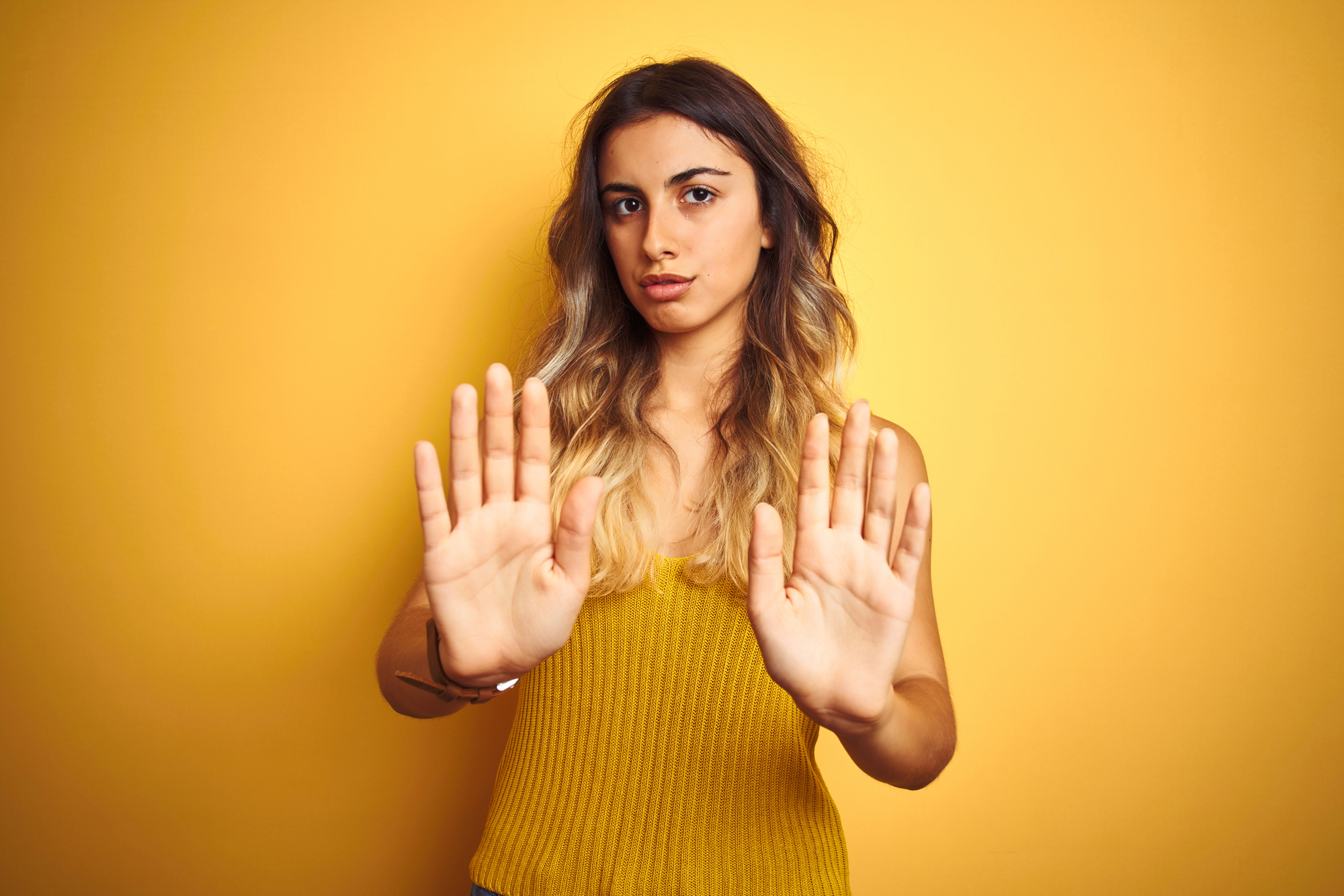 A woman with long, wavy hair stands against a yellow background, wearing a yellow sleeveless top. She has a serious expression and is holding both hands up with palms facing forward in a stop gesture.