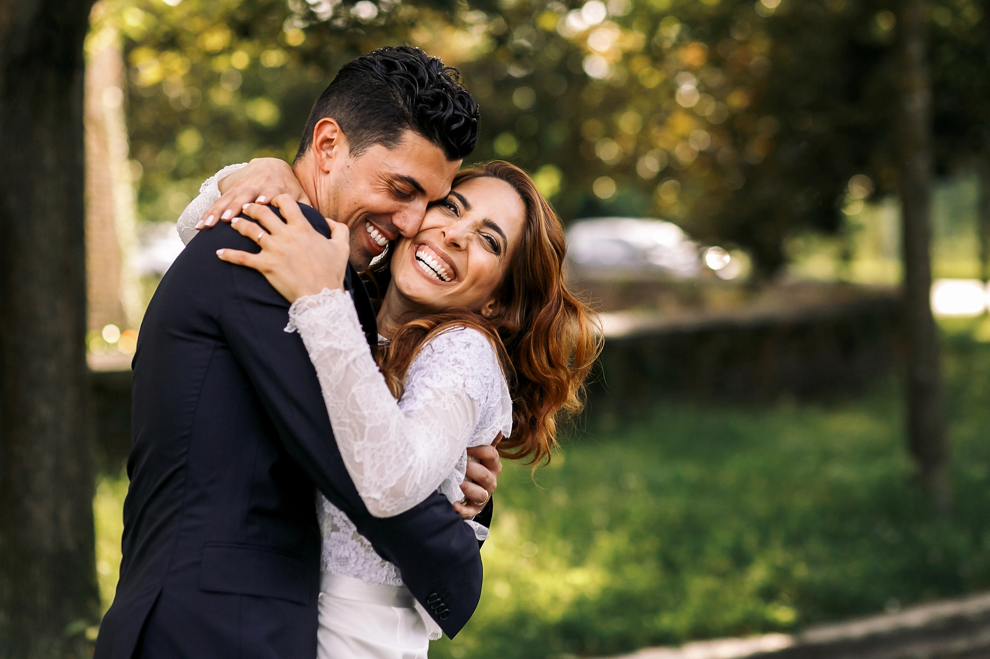 A smiling couple embraces in a joyful hug in a lush, green park. The man is wearing a dark suit, and the woman is dressed in a white lace garment. Both look happy and content, with sunlight filtering through the trees in the background.