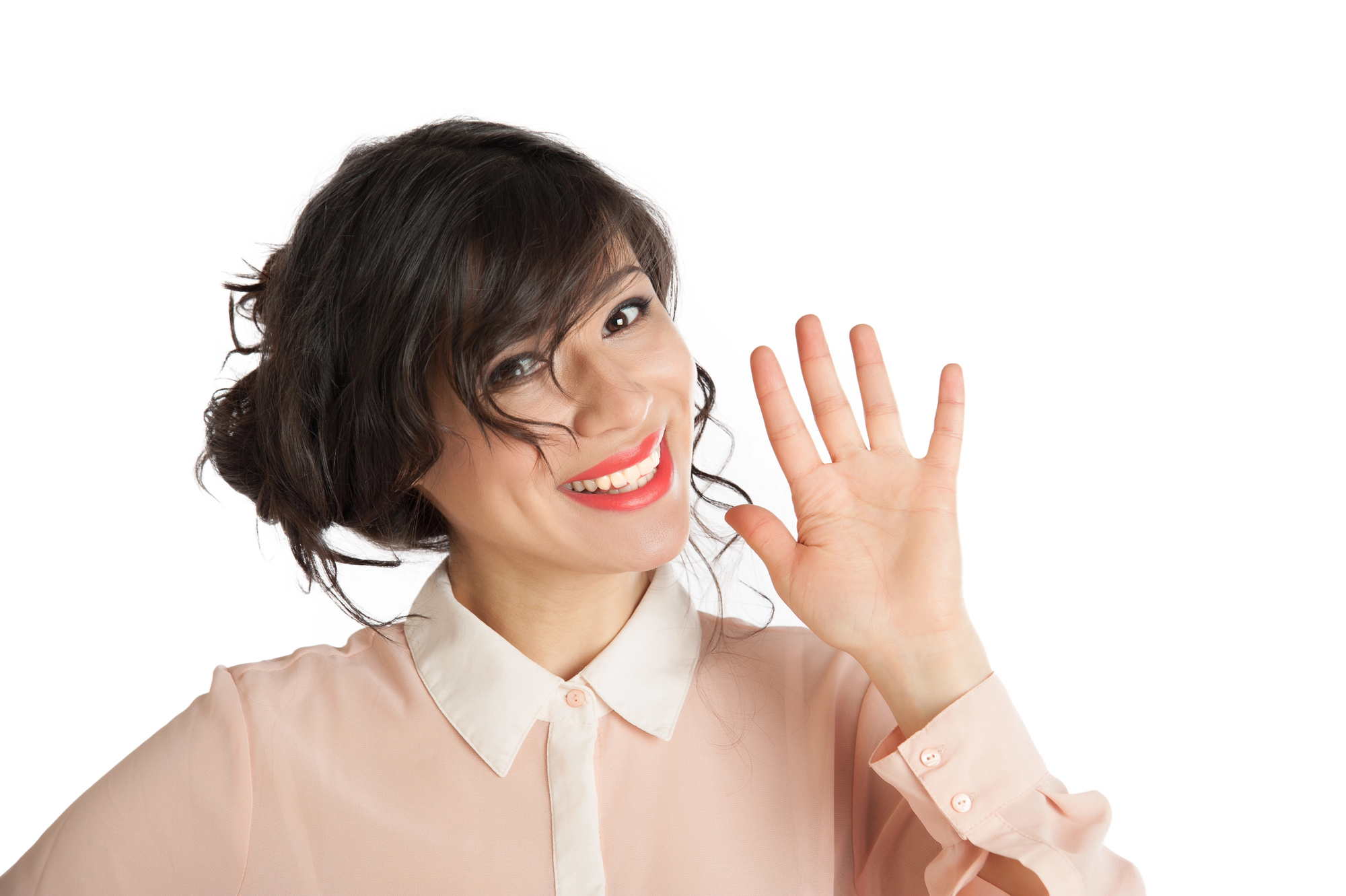 A smiling woman with dark hair in a loosely tied updo, wearing a light pink blouse with a white collar, waves at the camera. She stands against a plain white background.