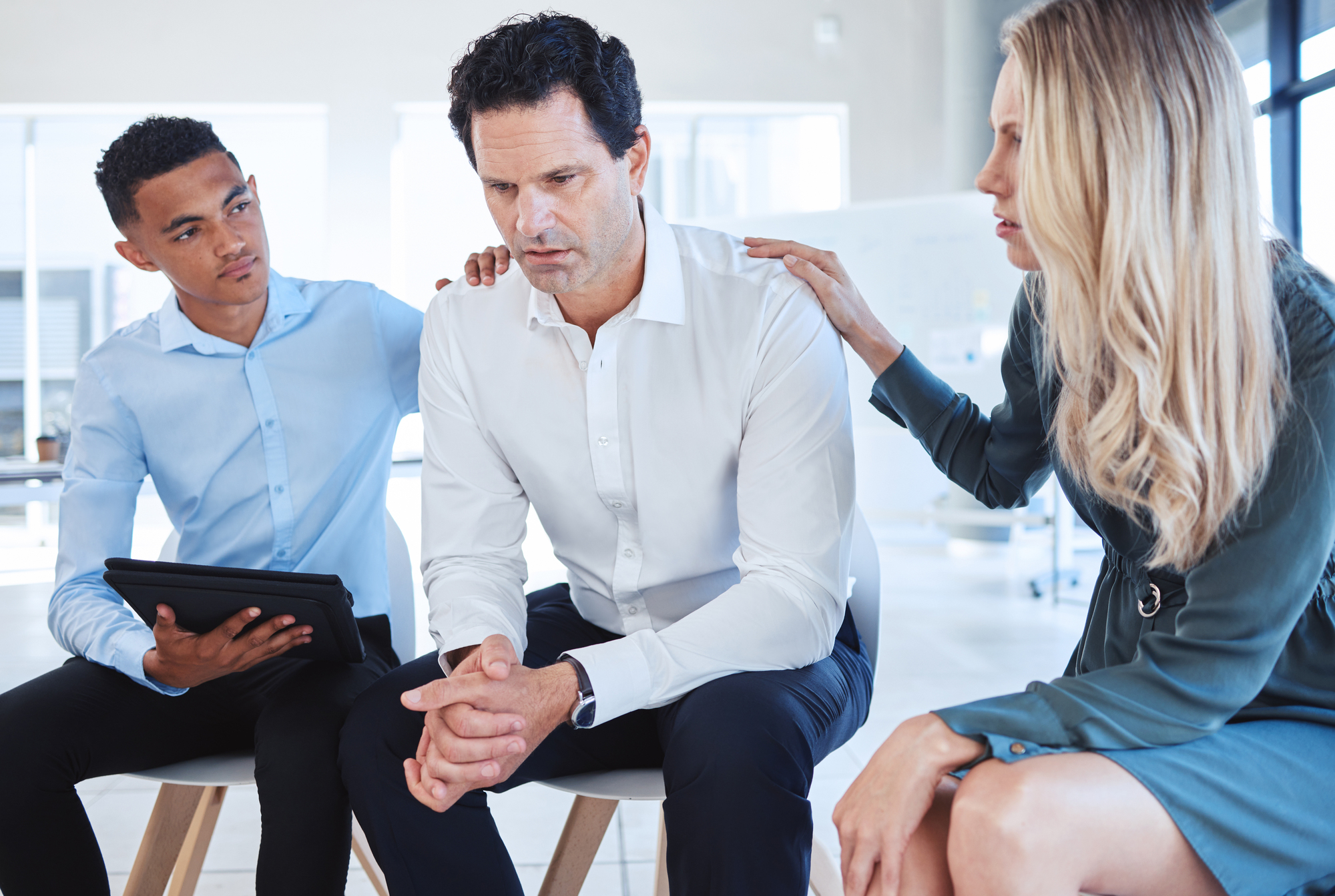 A worried man in a white shirt sits on a chair, leaning forward with his hands clasped. A young man in a light blue shirt holds a tablet and places his hand on the man's shoulder. A woman in a green dress also comforts the man with her hand on his other shoulder.