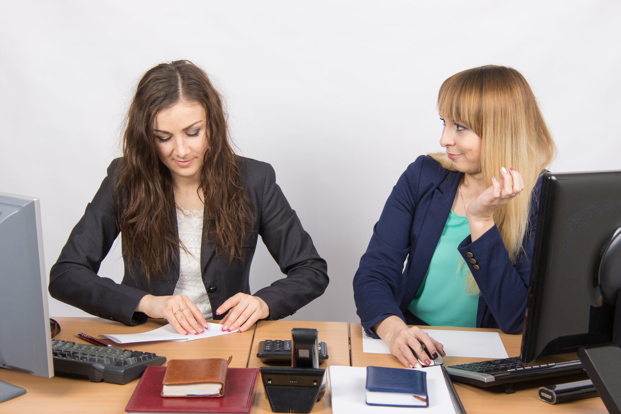 Two women are seated at a desk in an office setting. The woman on the left is focused on paperwork, while the woman on the right, seated beside a computer, appears to be glancing at her colleague. The desk is cluttered with office supplies, notebooks, and a calculator.