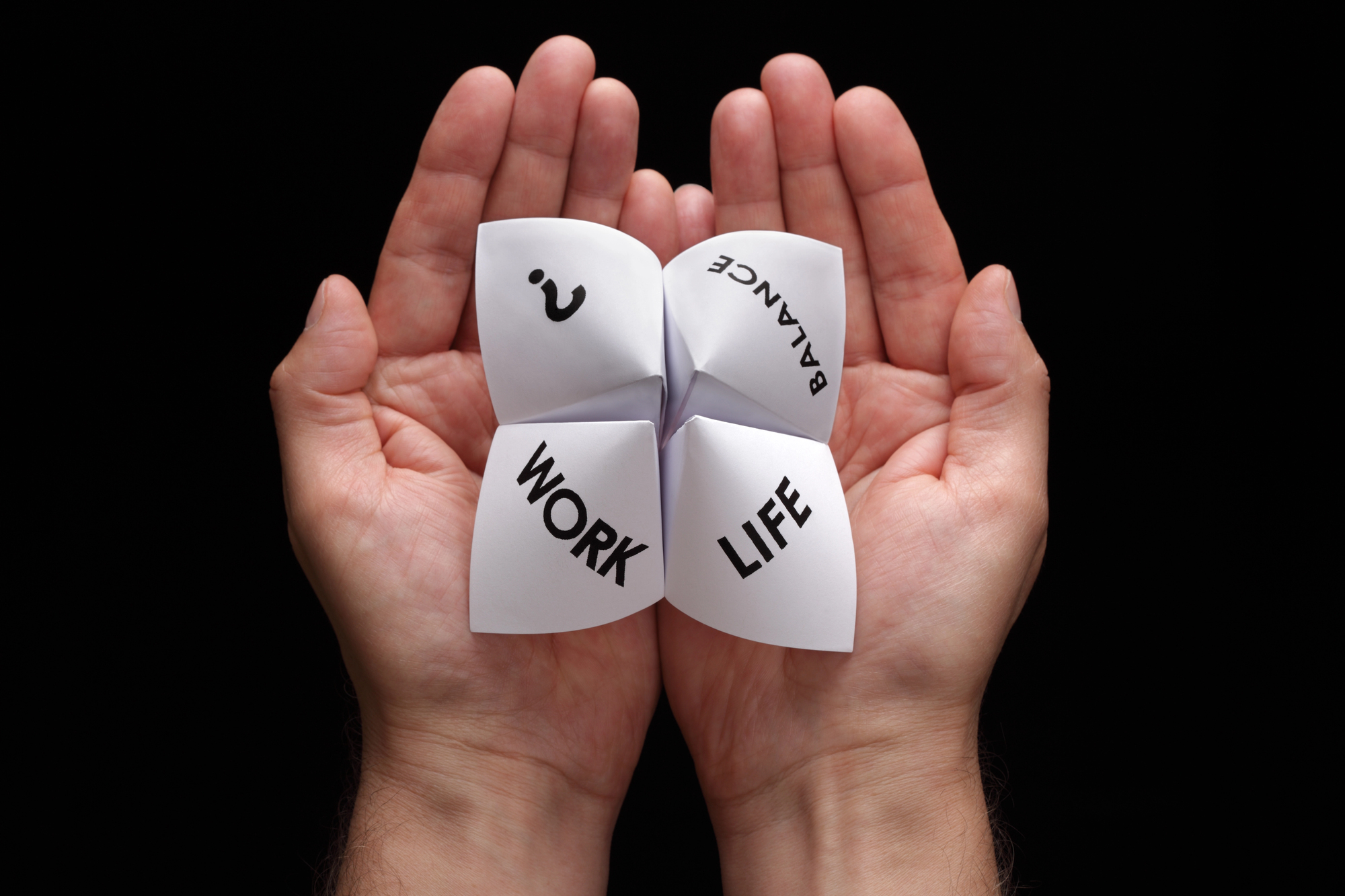 A pair of hands holds a white paper fortune teller with four visible sections labeled "Work," "Life," "Balance," and a question mark. The background is black, making the hands and paper fortune teller stand out.