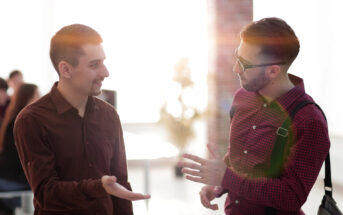 Two men are engaged in a conversation in a well-lit room with sunlight streaming through the window. One man wears a brown shirt, and the other wears a red checked shirt and glasses. People can be seen in the background blurry, possibly working or socializing.