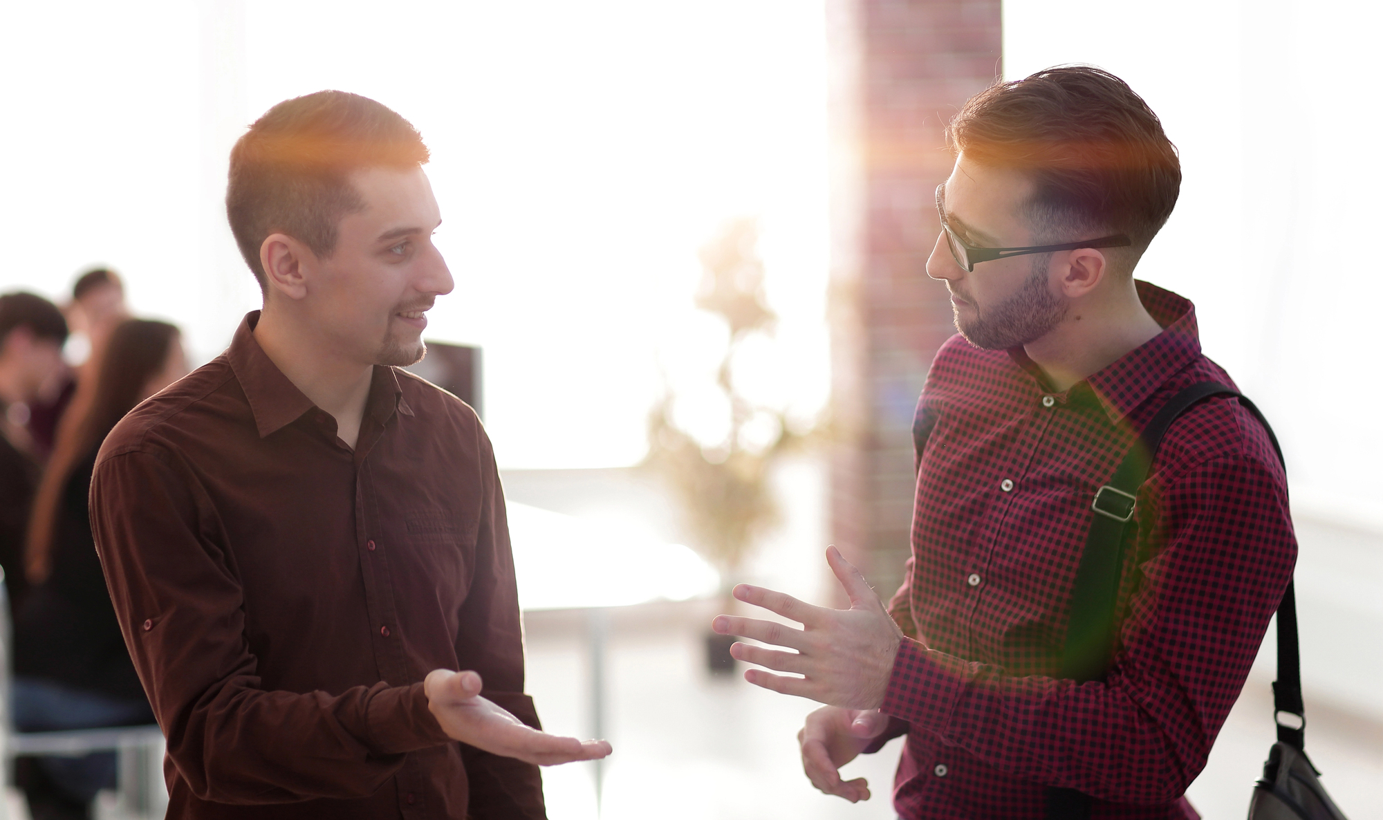 Two men are engaged in a conversation in a well-lit room with sunlight streaming through the window. One man wears a brown shirt, and the other wears a red checked shirt and glasses. People can be seen in the background blurry, possibly working or socializing.