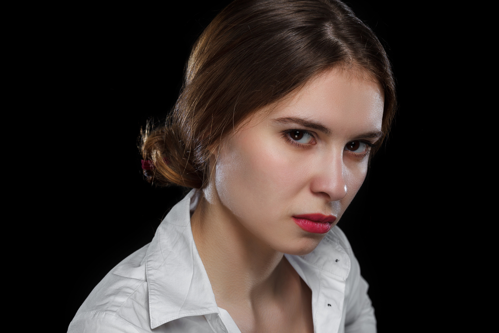 A woman with light skin and long brown hair pulled back into a low bun is wearing a white collared shirt. She has a neutral expression and is looking directly at the camera against a solid black background.