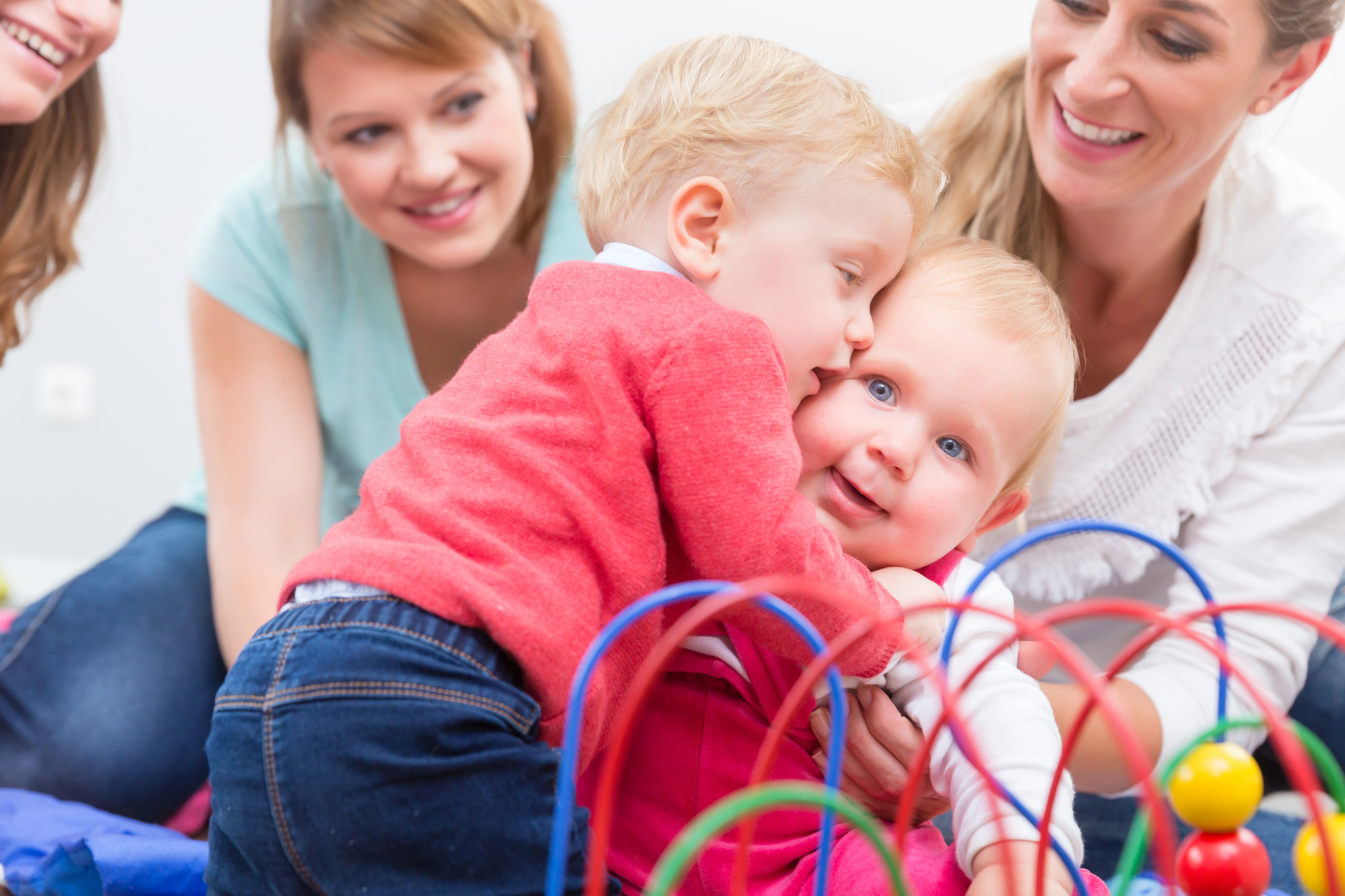 Two toddlers, one in a red sweater and one in a pink outfit, interact and hug while playing with a colorful wire bead maze toy. Two women, smiling, watch the toddlers affectionately from the background. The scene is bright and cheerful.