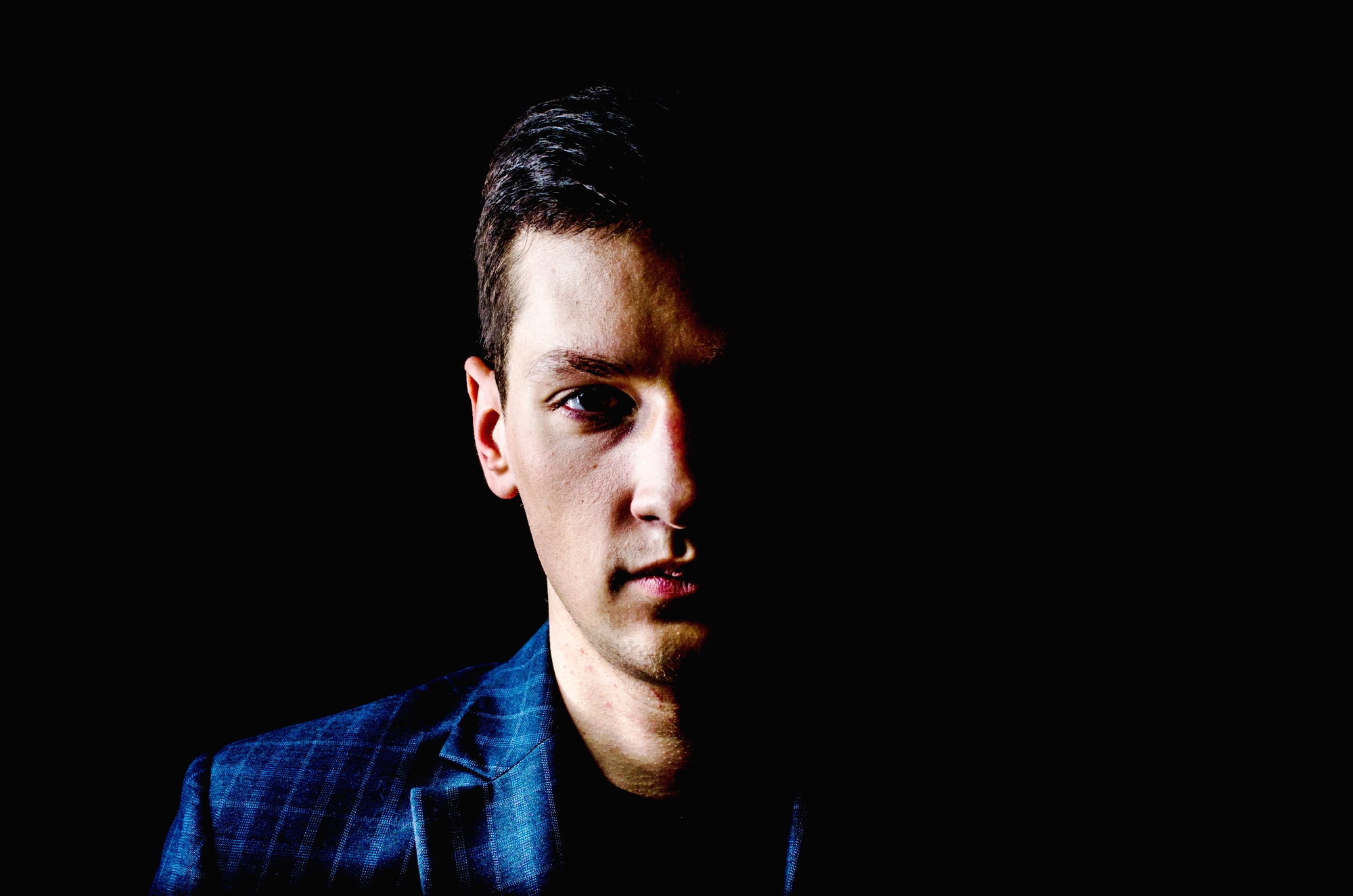 A close-up portrait of a man against a dark background. Only half of his face is illuminated, highlighting his serious expression while the other half is in shadow. He is wearing a dark blue checkered blazer.