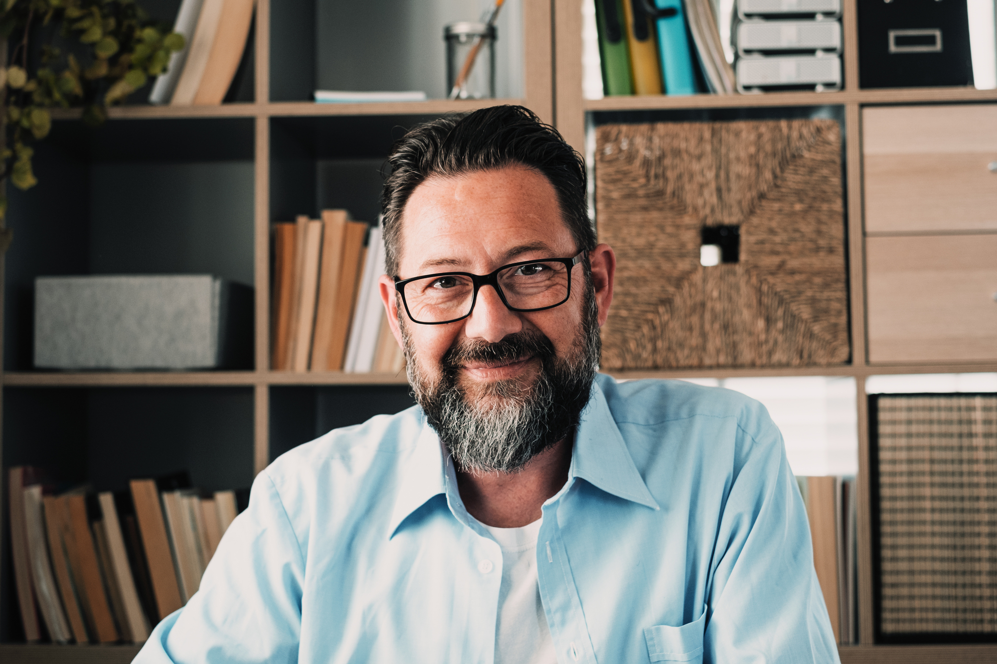 A bearded man with glasses, wearing a light blue shirt, smiles while seated in front of a bookshelf filled with books and boxes. The background features a variety of colorful files and office supplies.