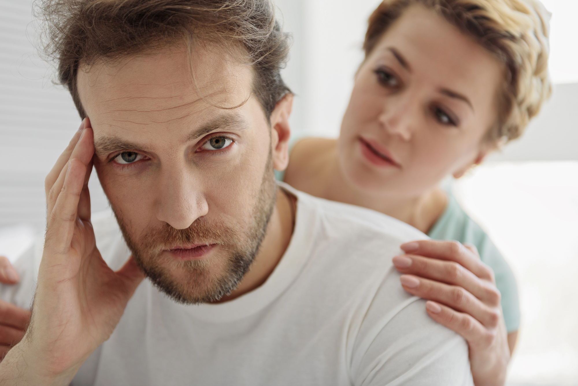 A man with a worried expression rests his head on one hand while a woman stands behind him with a concerned look, gently placing her hand on his shoulder. They are in a bright room and both appear to be in a serious conversation.