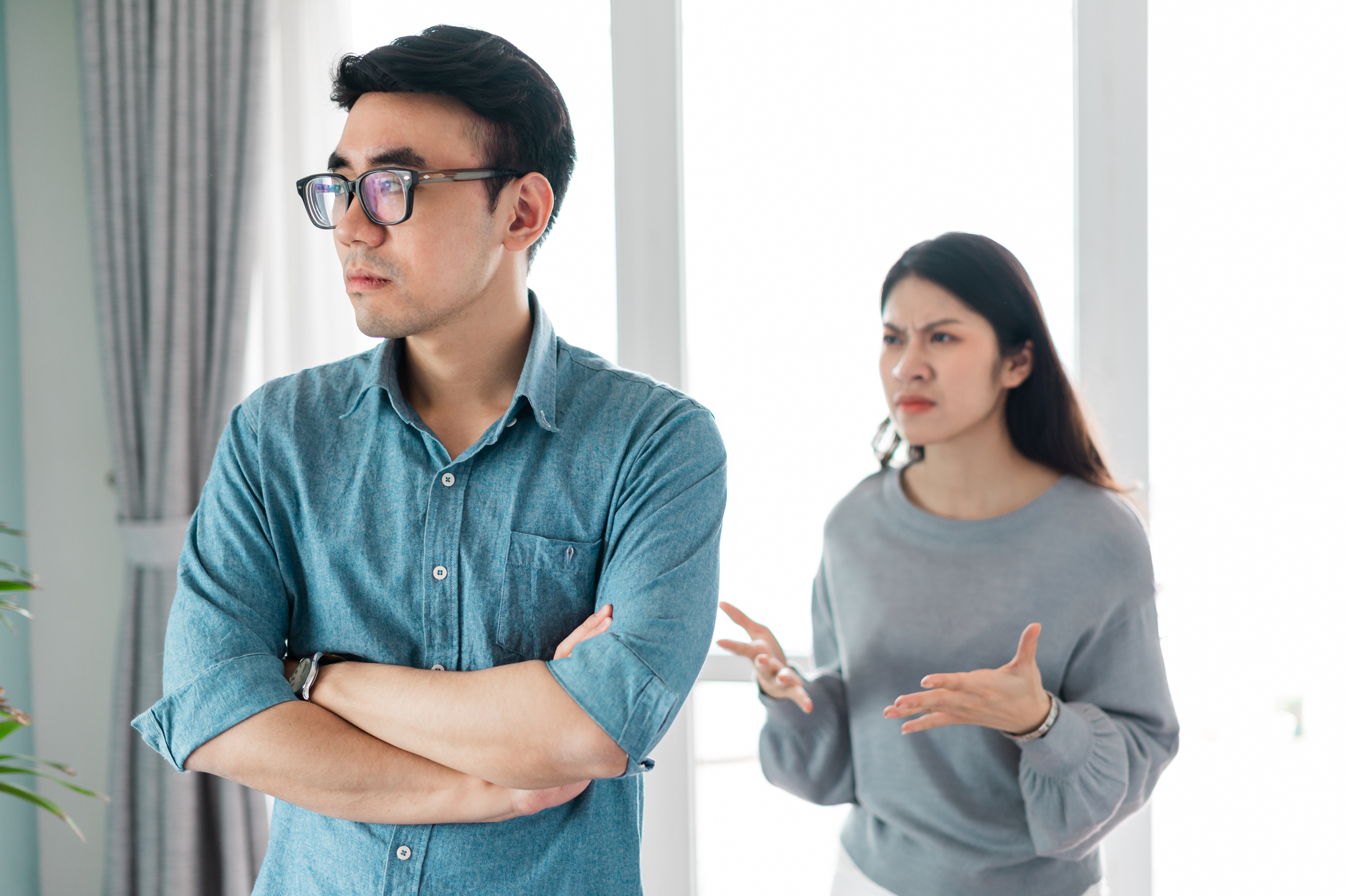 A man with glasses, wearing a blue shirt, stands with his arms crossed and looks away. A woman with long hair and a gray sweater stands behind him, with her arms out and a frustrated expression. They are in a bright room with curtains and large windows.