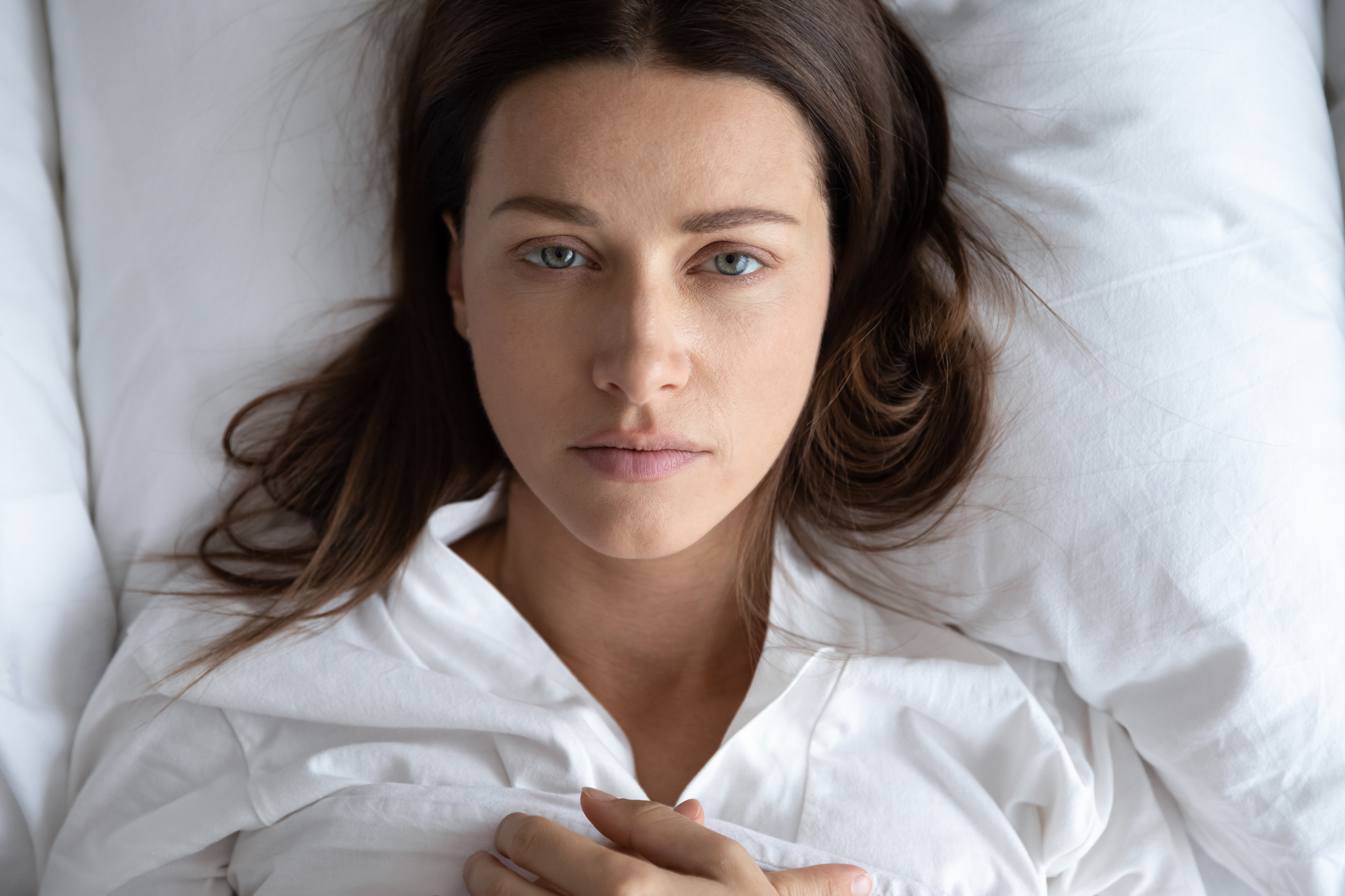 A woman with long brown hair lies on a bed, staring directly at the camera. She is dressed in a white shirt and surrounded by white pillows, with a calm expression on her face. The overall atmosphere is serene and relaxed.