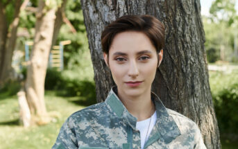 A person with short brown hair is leaning against a tree, wearing a camouflage-patterned military uniform. The background is lush with green trees and foliage, suggesting a park or outdoor setting. The individual is looking directly at the camera with a neutral expression.